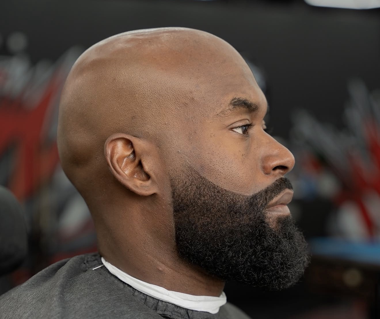 Side profile of a bald man with a full beard, looking to the right, in a barber shop setting.