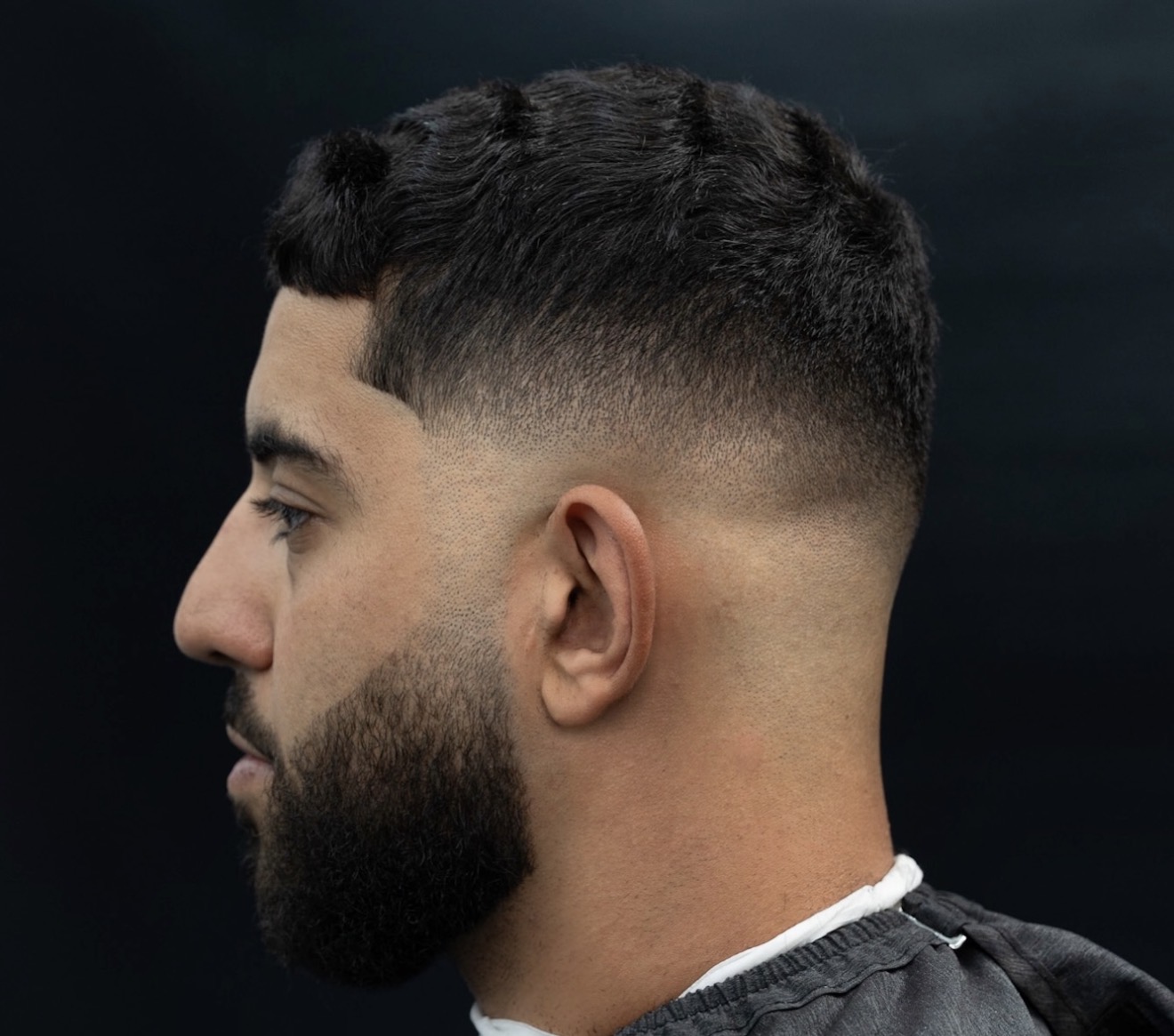 Side profile of a man with a short, styled haircut and beard, wearing a dark shirt, against a dark background.