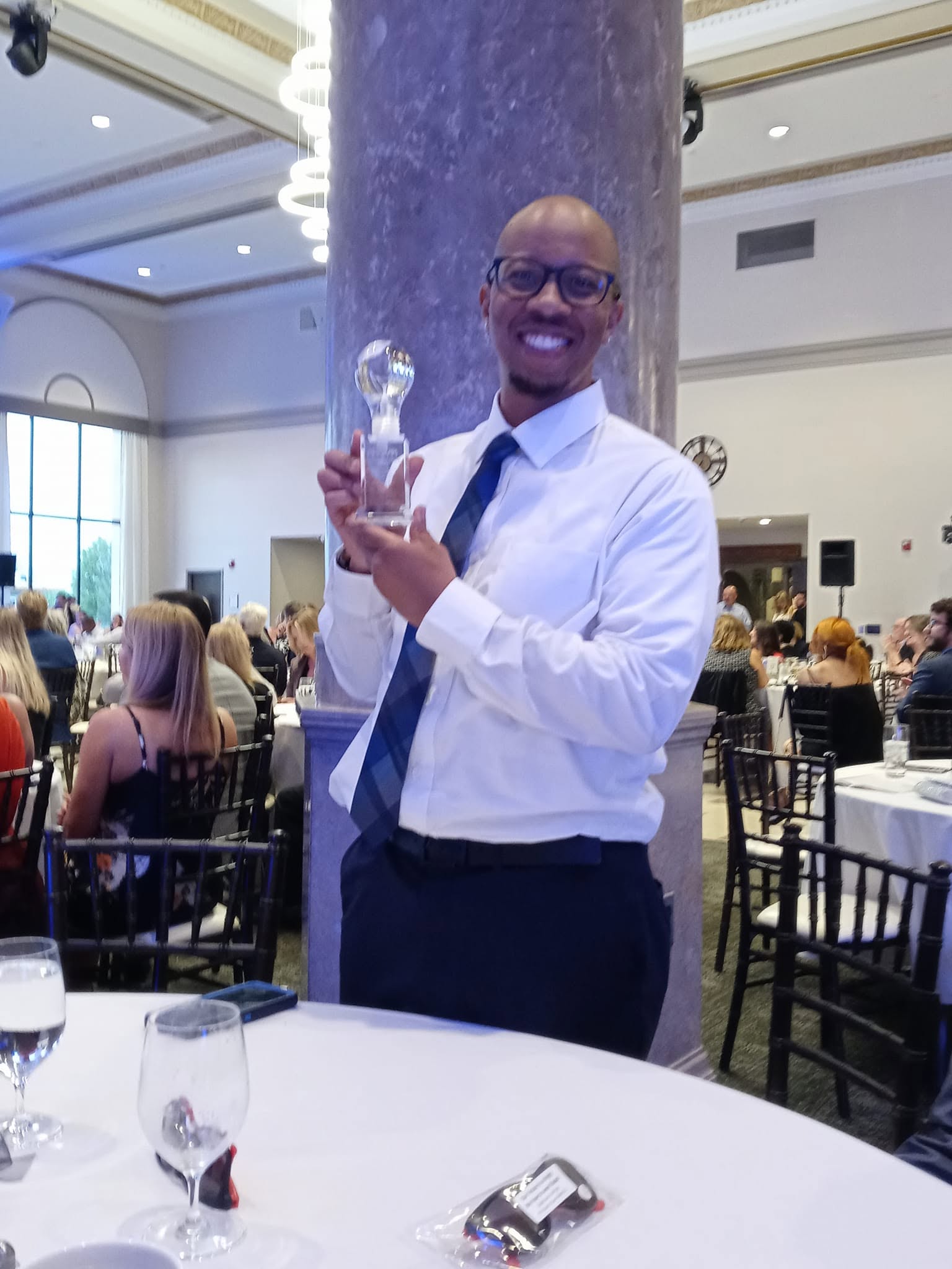 Man in white shirt and tie holding an award, smiling in a banquet hall with seated guests and tables.