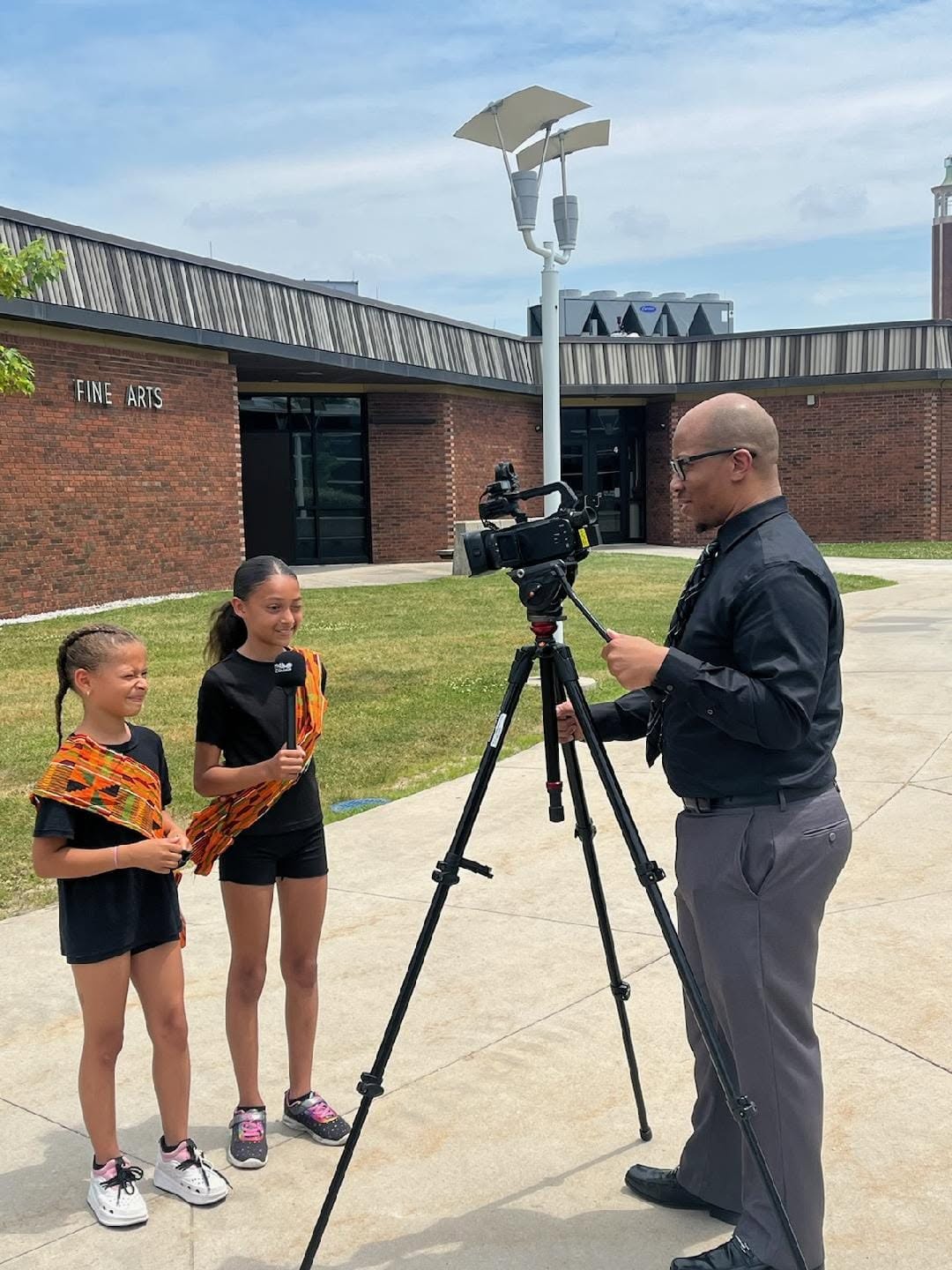 Two young girls being interviewed by a man with a camera outdoors on a sunny day, brick building in background.