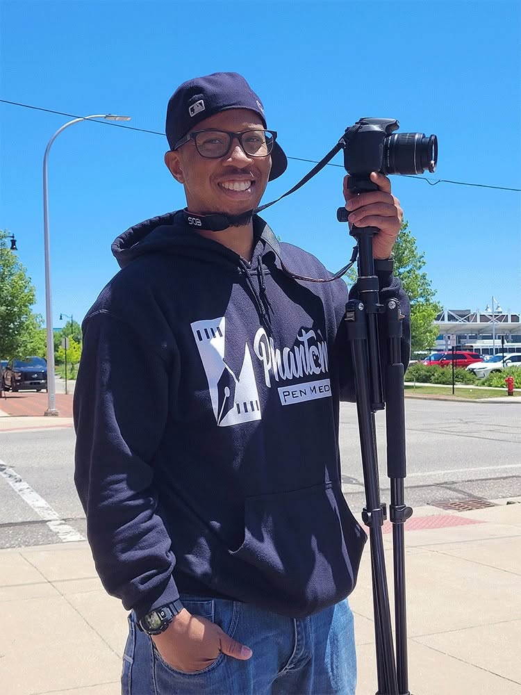Person smiling, wearing glasses and a hoodie, holding a camera on a tripod outdoors on a sunny day.