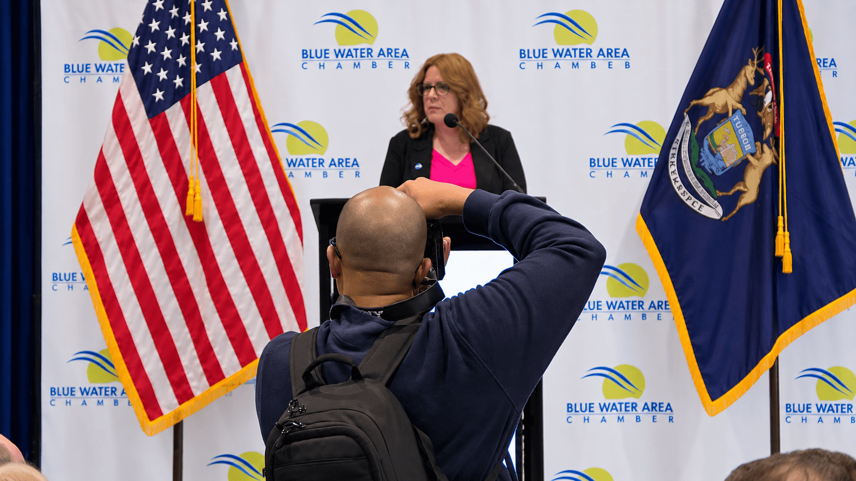 Woman speaking at a podium with American and state flags, Blue Water Area backdrop, audience member taking photo.