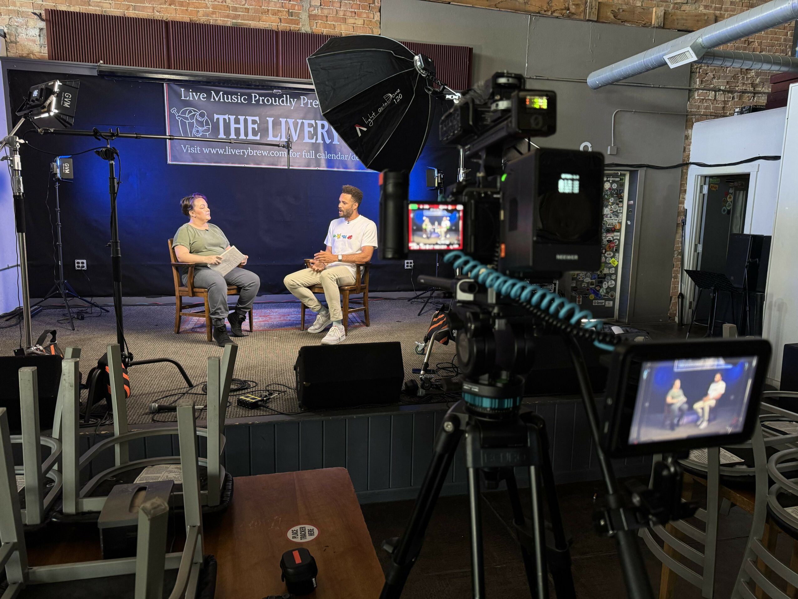 Two people seated on stage with microphones, a camera recording the scene, and a backdrop reading 'The Liver'.