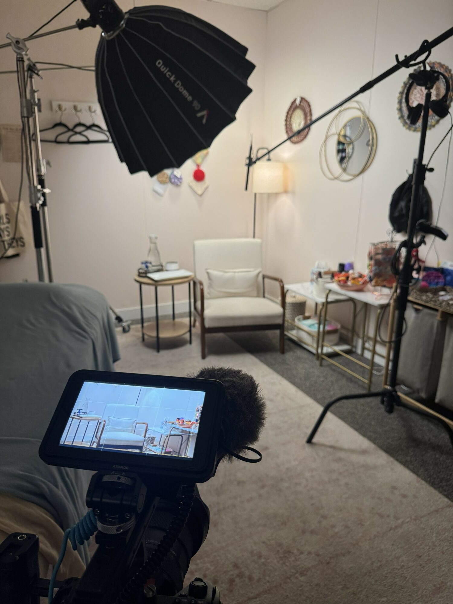 Indoor filming setup with lighting, camera, and a white chair in a room with wall decor and a side table.