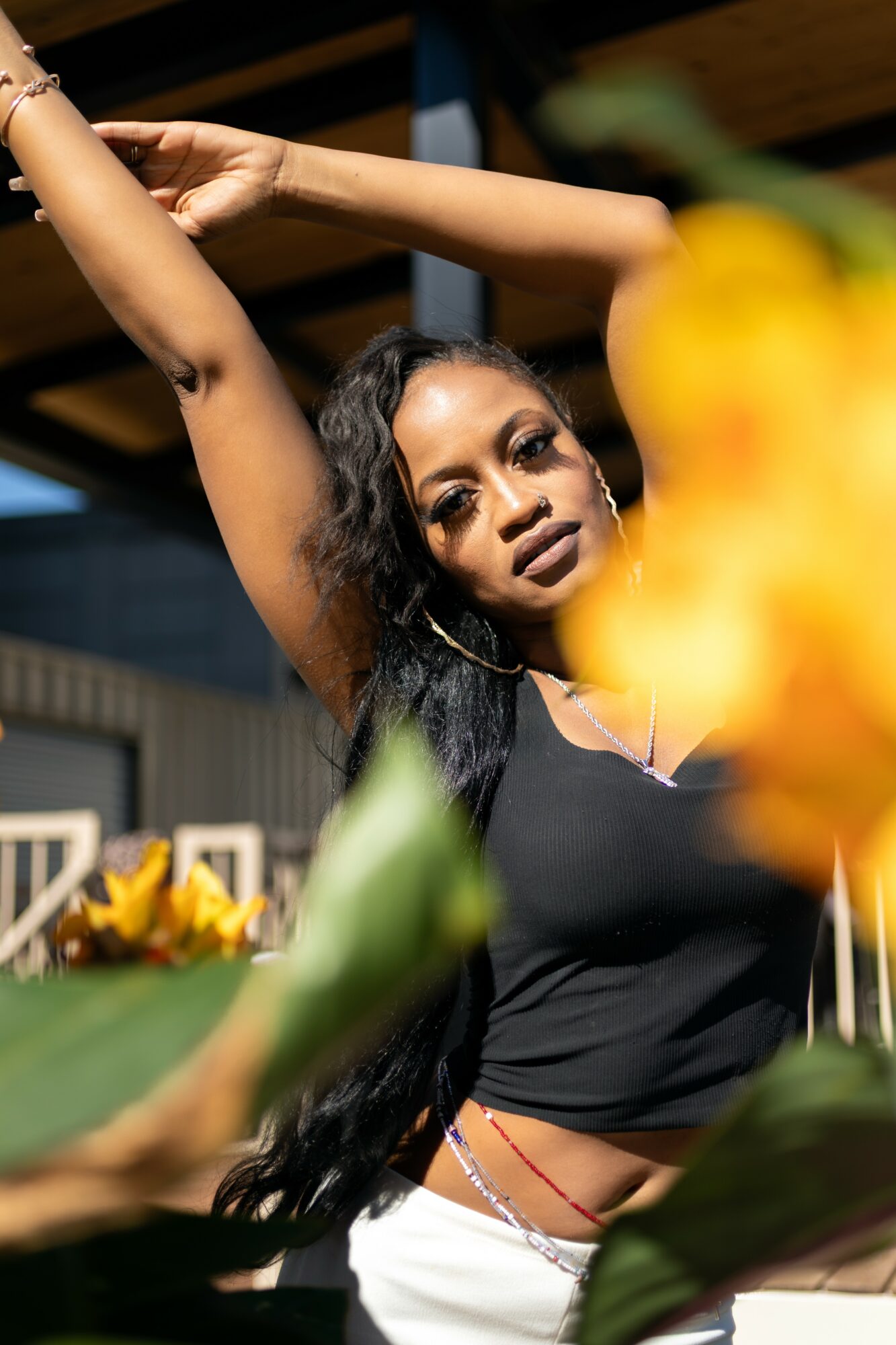 Woman with long dark hair and dark lipstick poses with arms raised, surrounded by colorful flowers, indoors.