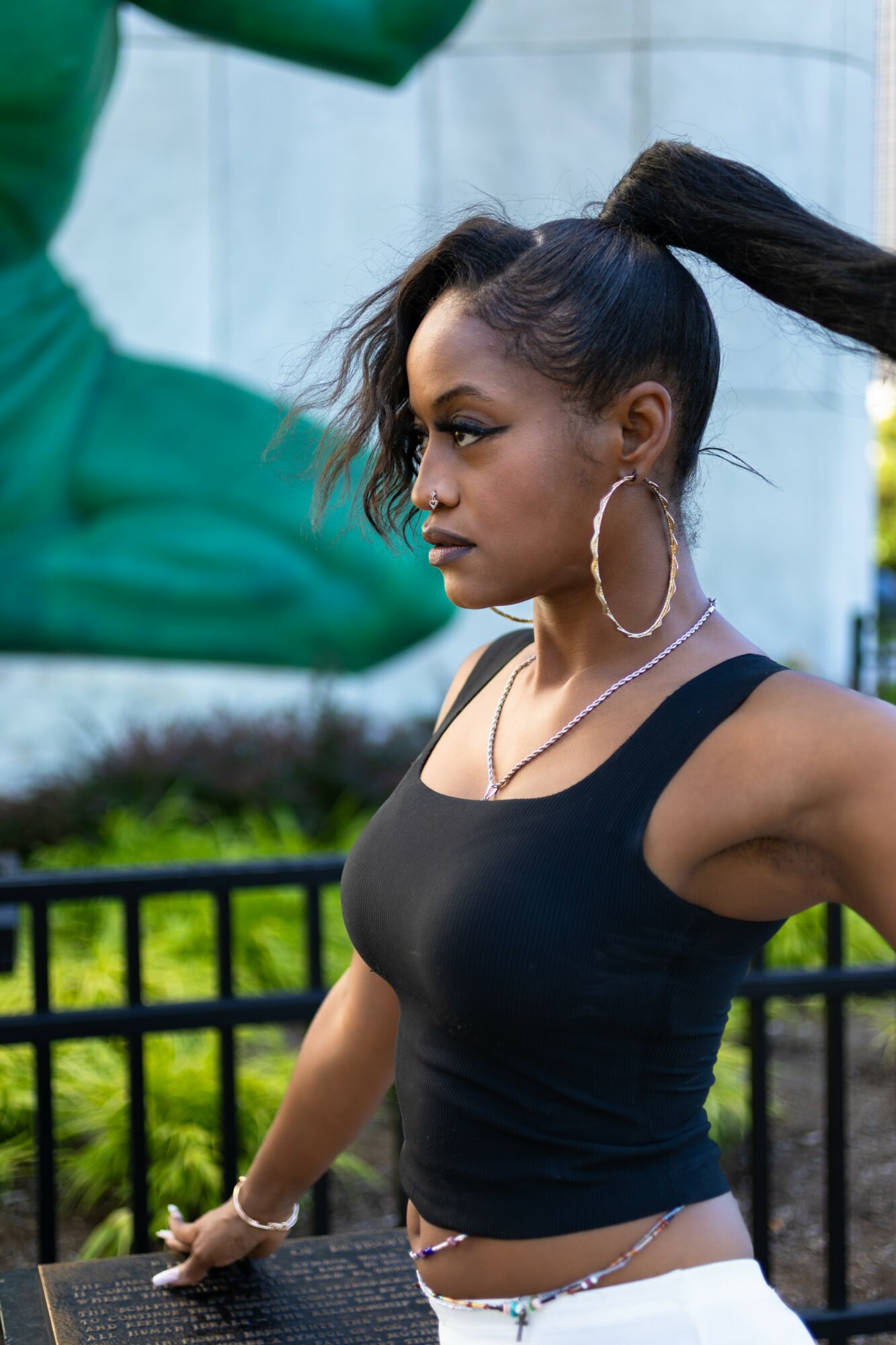 Young woman with long hair in a high ponytail, wearing hoop earrings and a black top, outdoors near a statue.