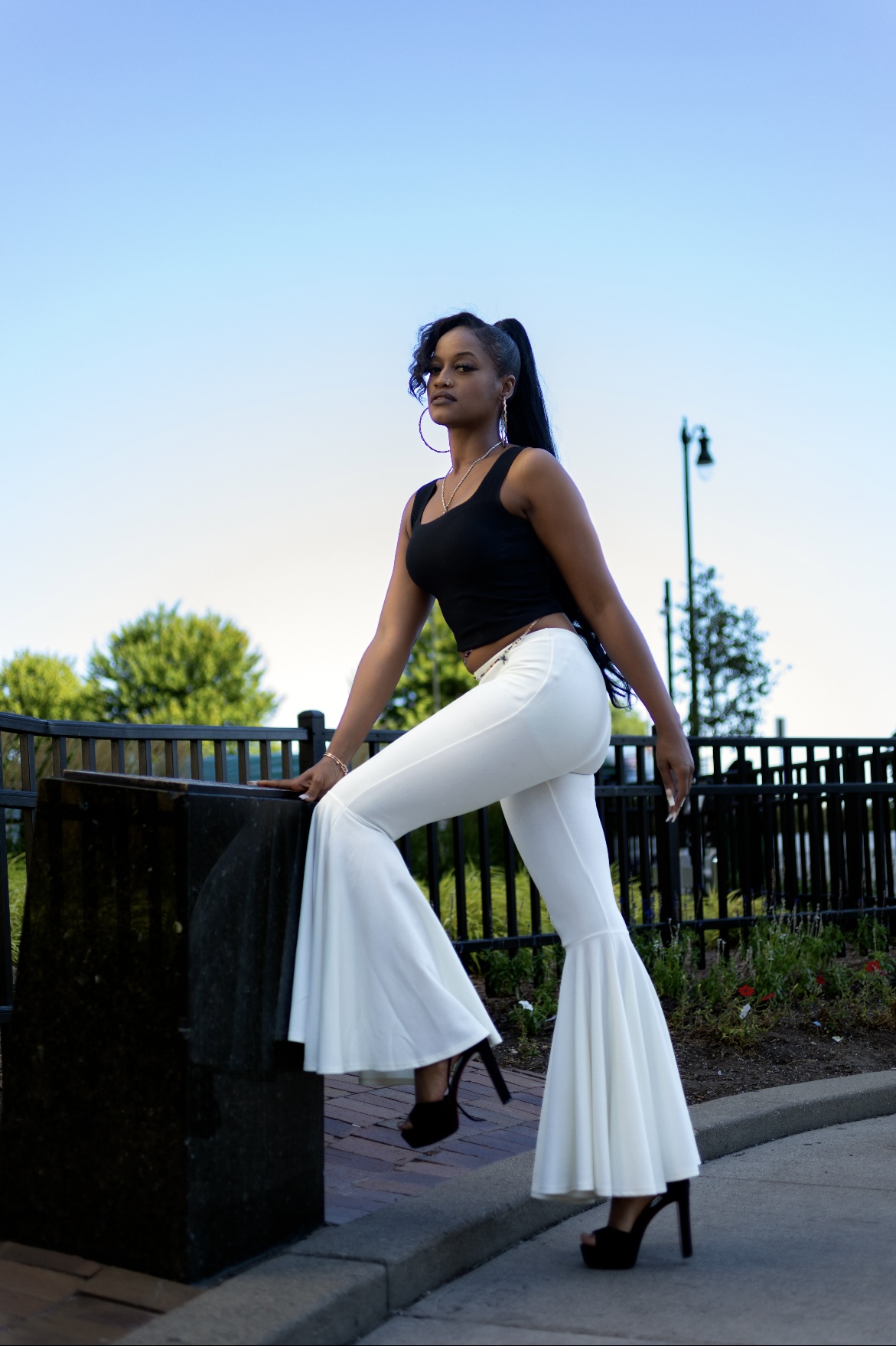 Woman in black top and white flared pants standing outdoors near a railing, with trees and a lamppost in background.