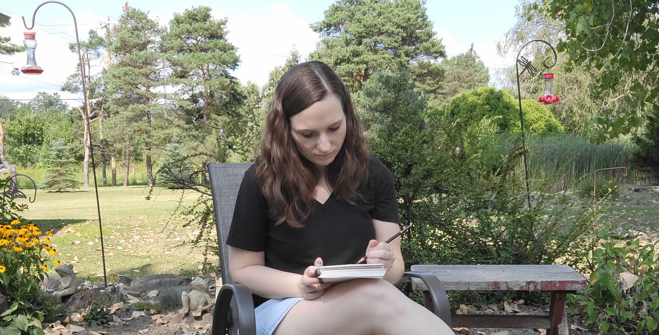 Young woman sitting outdoors on a bench, writing in a notebook, surrounded by trees and plants.