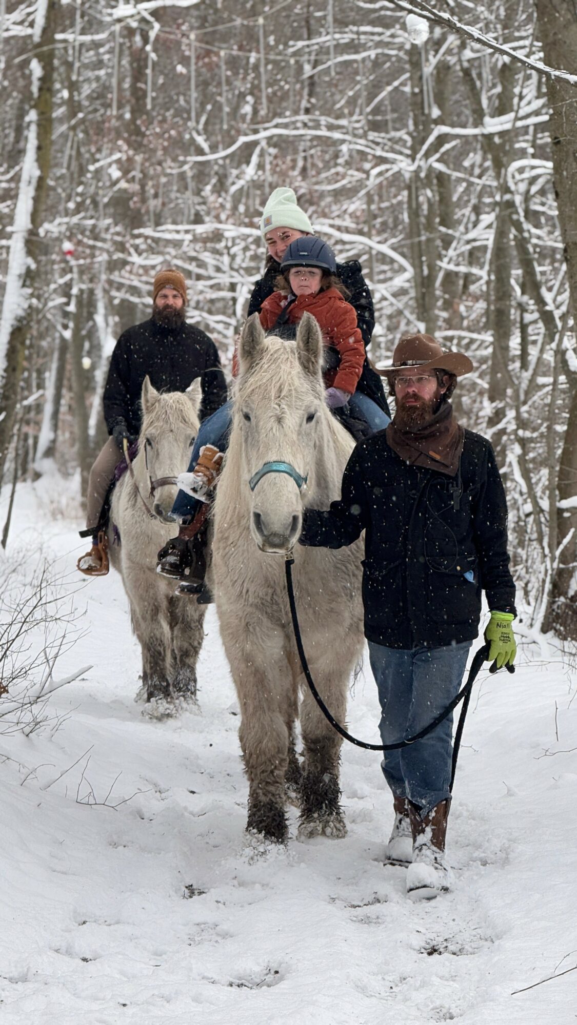 Group of people riding horses through snowy forest, led by a person holding a white horse's reins.