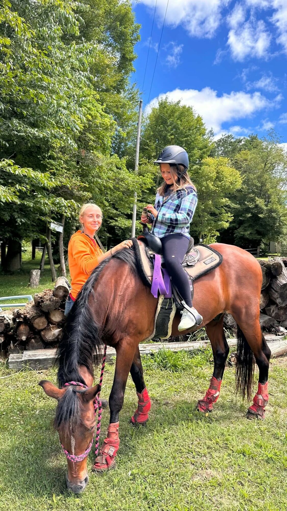 Girl in helmet riding a brown horse with purple saddle and red leg wraps, grazing on grass, with a woman nearby, trees, and blue sky.
