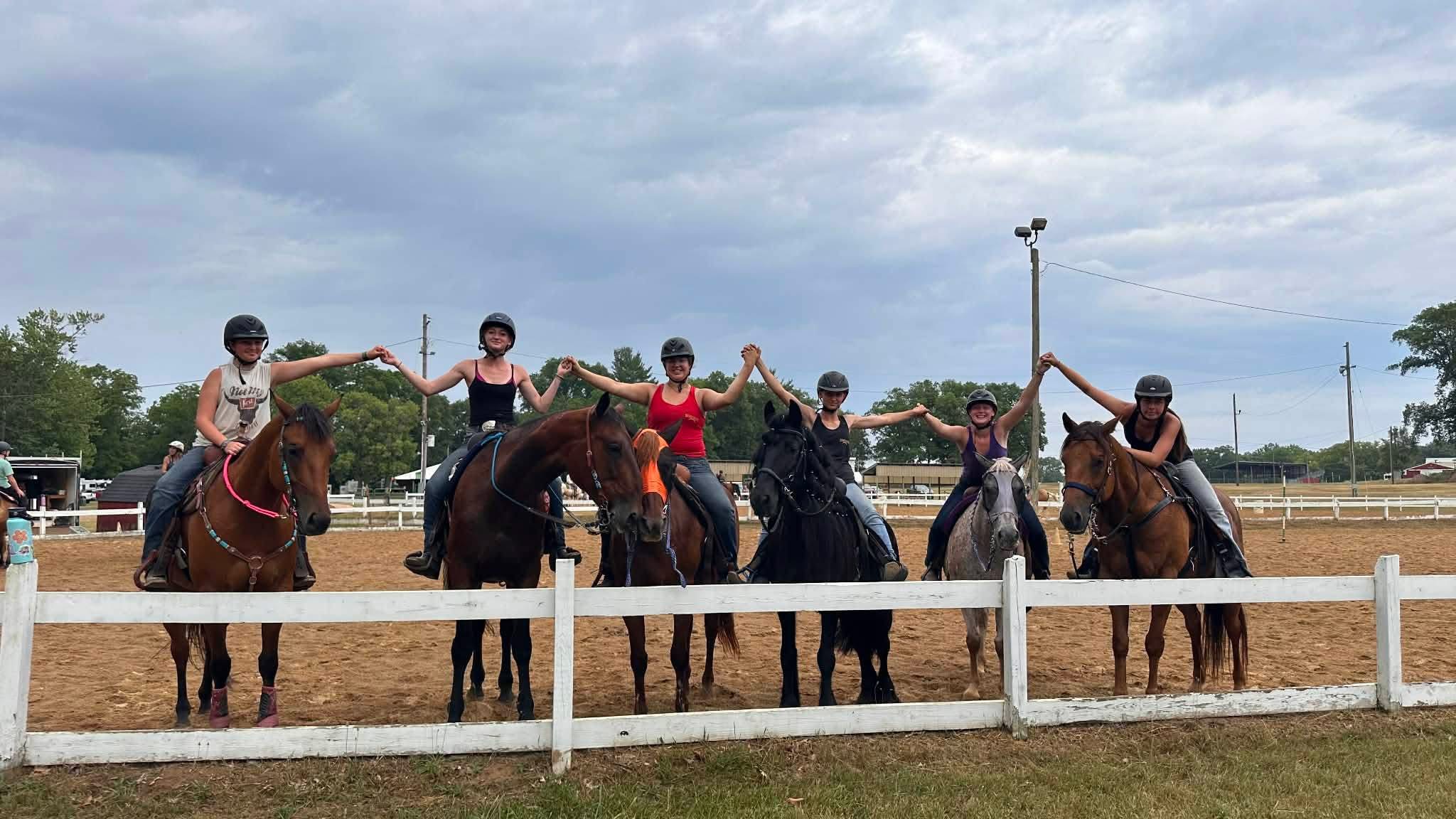 Six people on horseback holding hands in a line at an outdoor riding arena under cloudy sky.