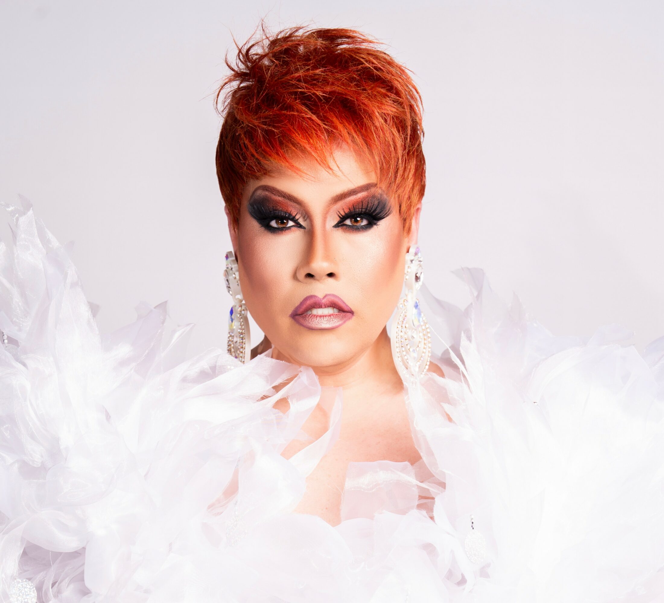 Woman with short red hair, dramatic makeup, and large earrings, wearing a white feathered outfit against a plain background.