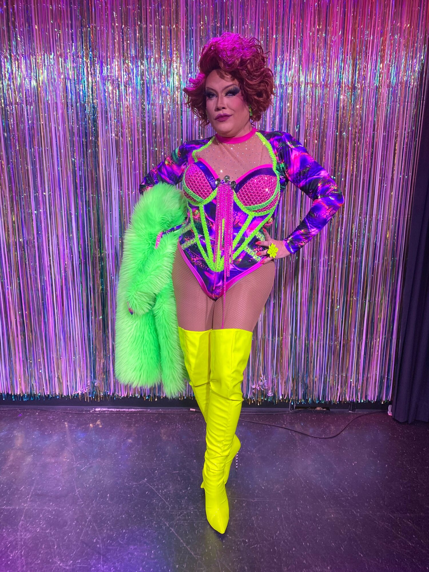 Person in colorful costume with neon yellow boots, purple and green accessories, standing in front of shiny curtain