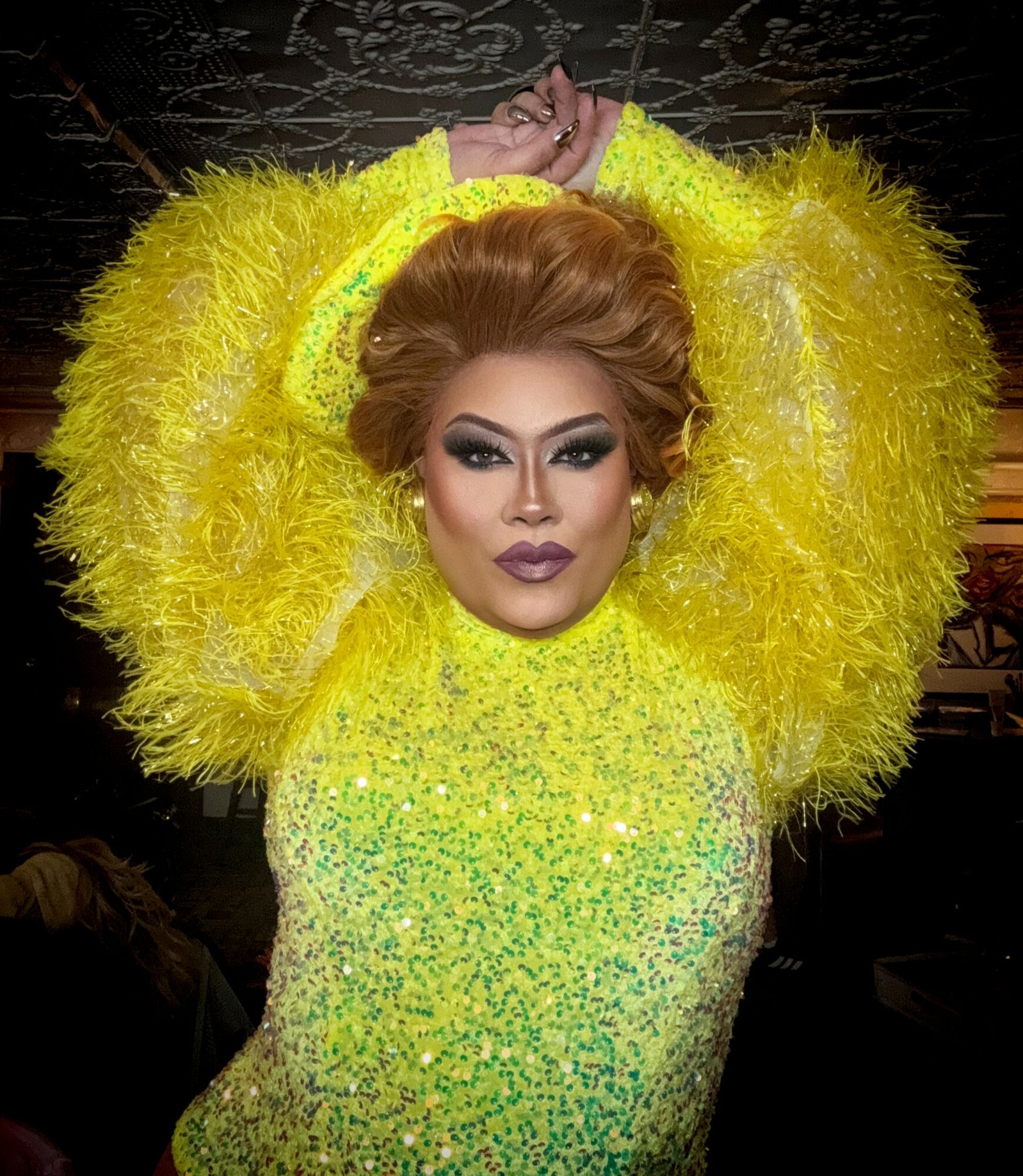 Person with styled hair and makeup, wearing a bright yellow, fluffy, sequined outfit, arms raised above head, dark background.