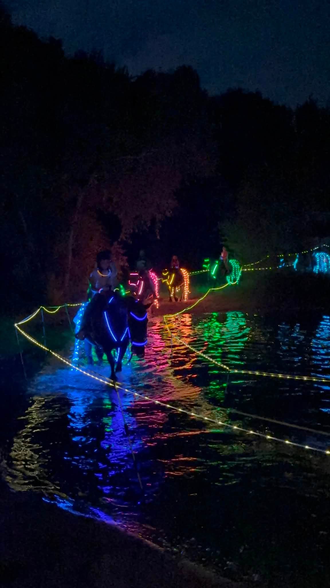 People riding horses with glowing neon lights along a river at night, reflecting in the water.