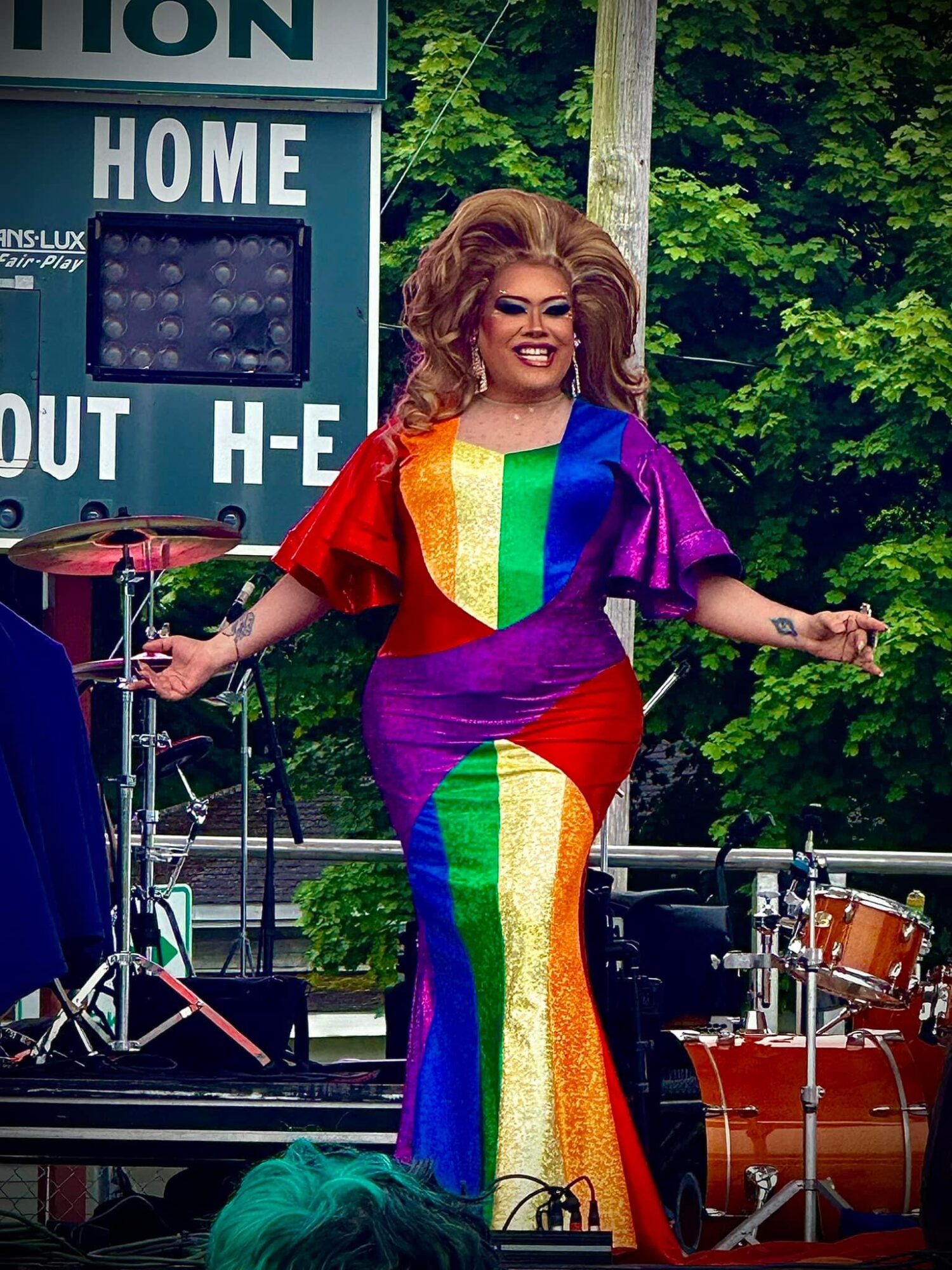 Person with curly hair in a colorful rainbow dress standing on an outdoor stage with musical instruments, smiling.