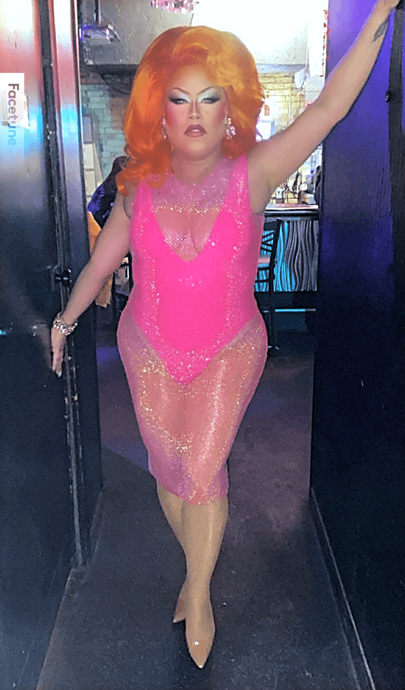 Person with bright orange hair in a pink, glittery dress walking through a doorway, arm raised, in a colorful setting.