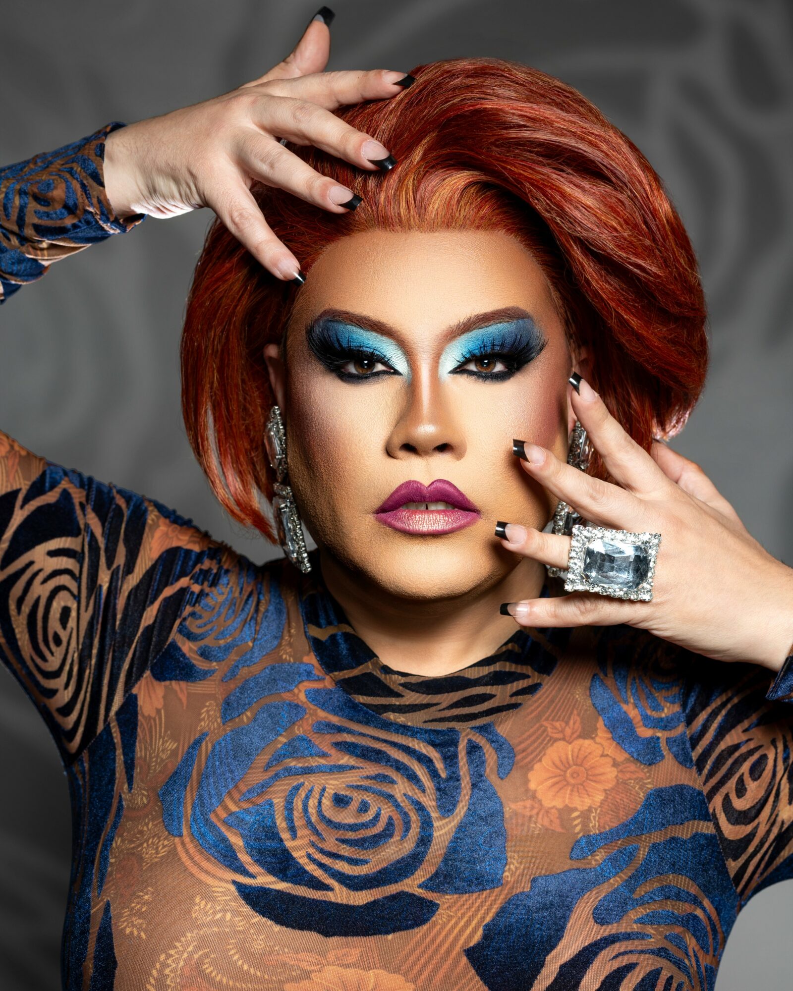 Woman with red hair, bold makeup, and large jewelry poses with hands near face against a gray background.