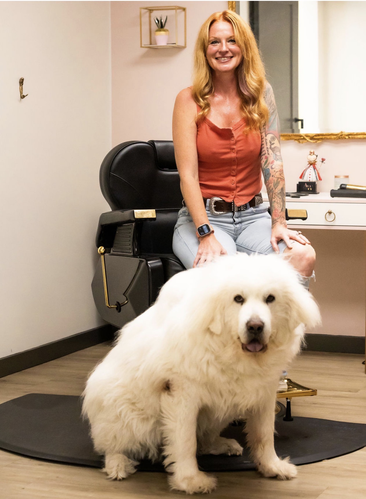Woman with long red hair smiling, sitting on a chair, with a large fluffy white dog in front, in a room with a sink.