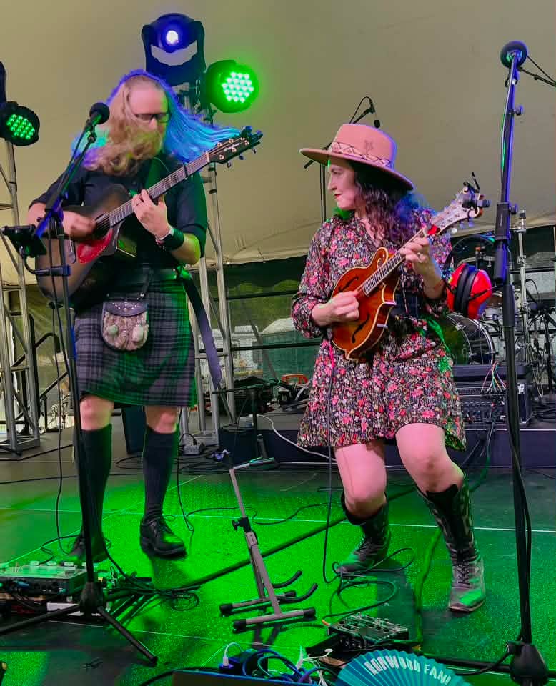 Two women playing guitars on stage with green and blue stage lights, wearing colorful clothing and hats, performing together.