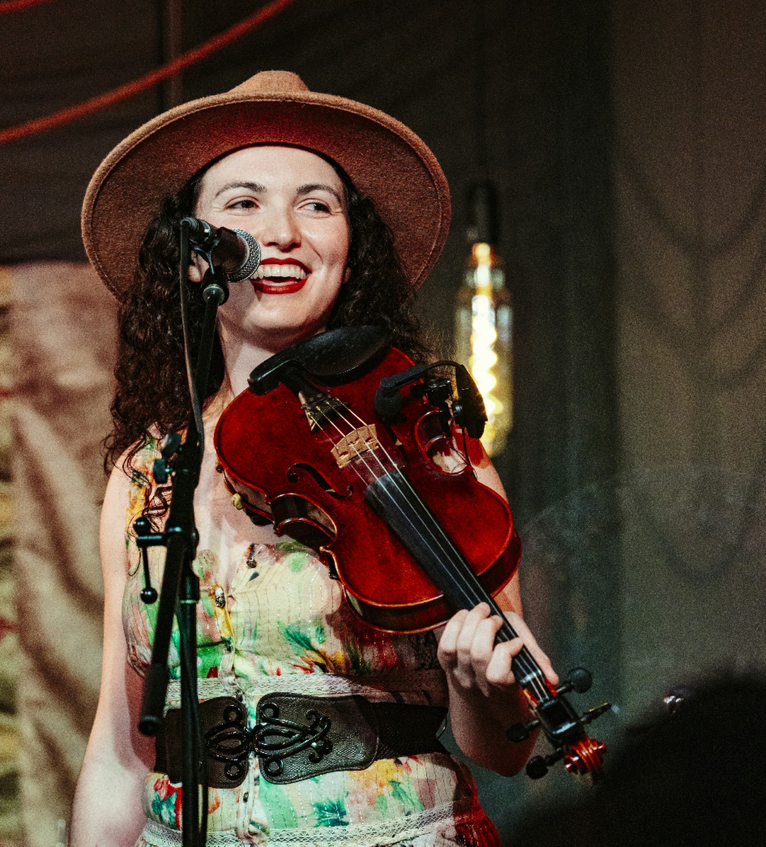 Woman with curly hair, wearing a wide-brimmed hat, holding a violin, smiling on stage with microphone, colorful background.