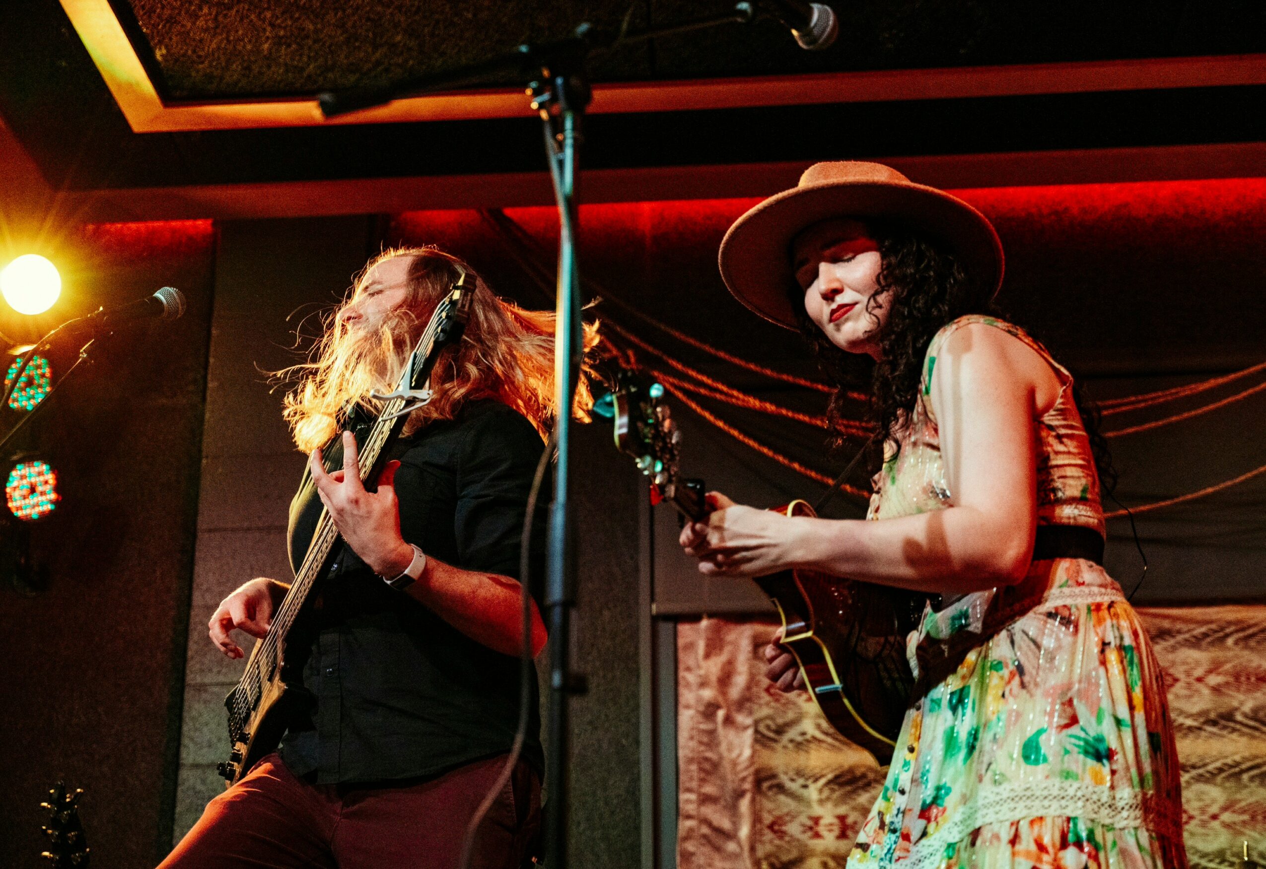 Two women playing guitars on stage with red lighting and a patterned backdrop, one wearing a hat and colorful skirt.