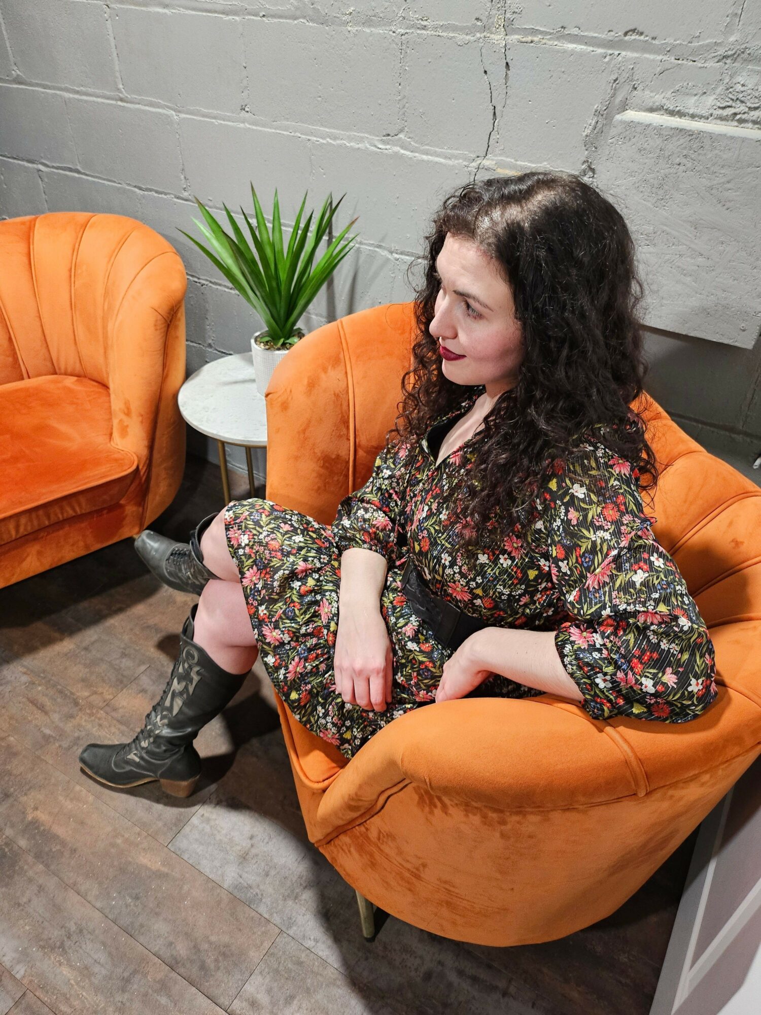 Woman with dark curly hair sitting in an orange chair, wearing a floral dress, in a room with a gray wall and wooden floor.