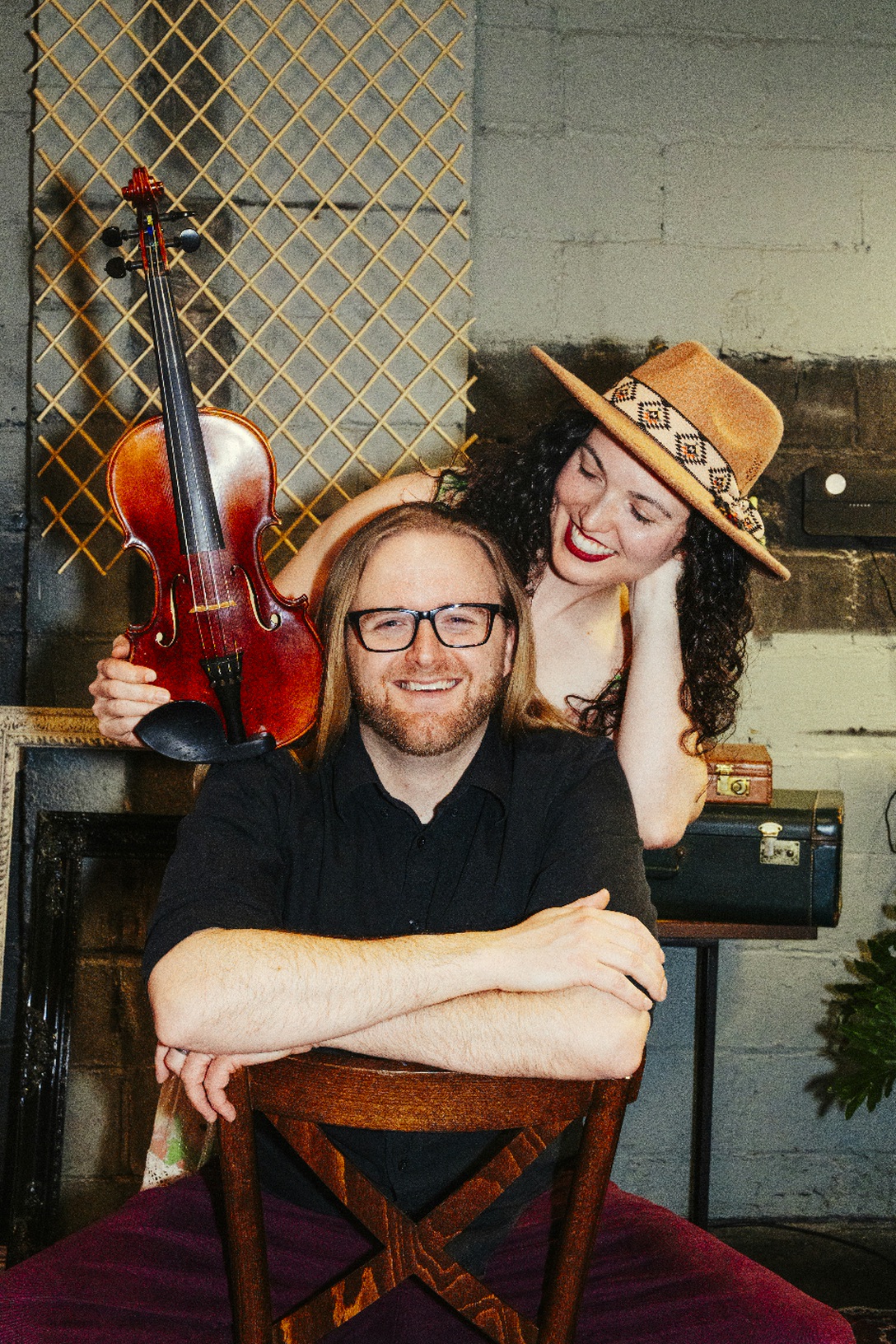 Two people smiling, one holding a violin, in a room with musical instruments and a lattice background.