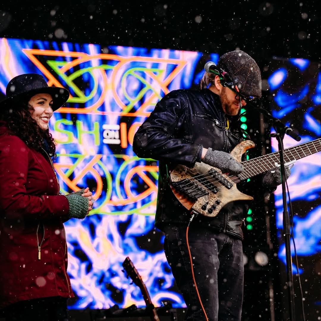 Two musicians perform on stage with a colorful background, one singing and the other playing a guitar, snow falling.