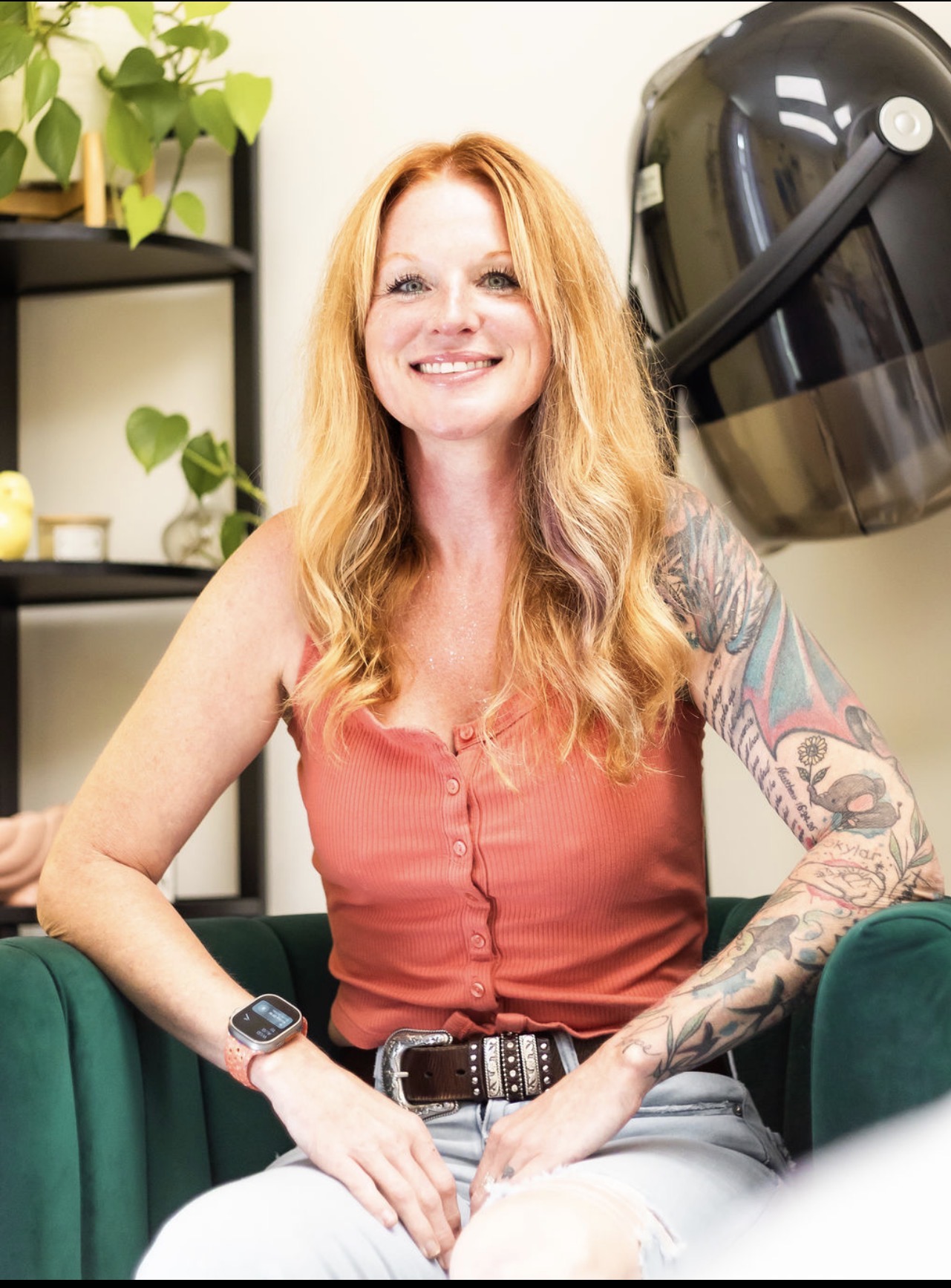 Woman with long red hair smiling, sitting in a salon, with a tattooed arm, wearing a sleeveless top, and a hairdryer nearby.
