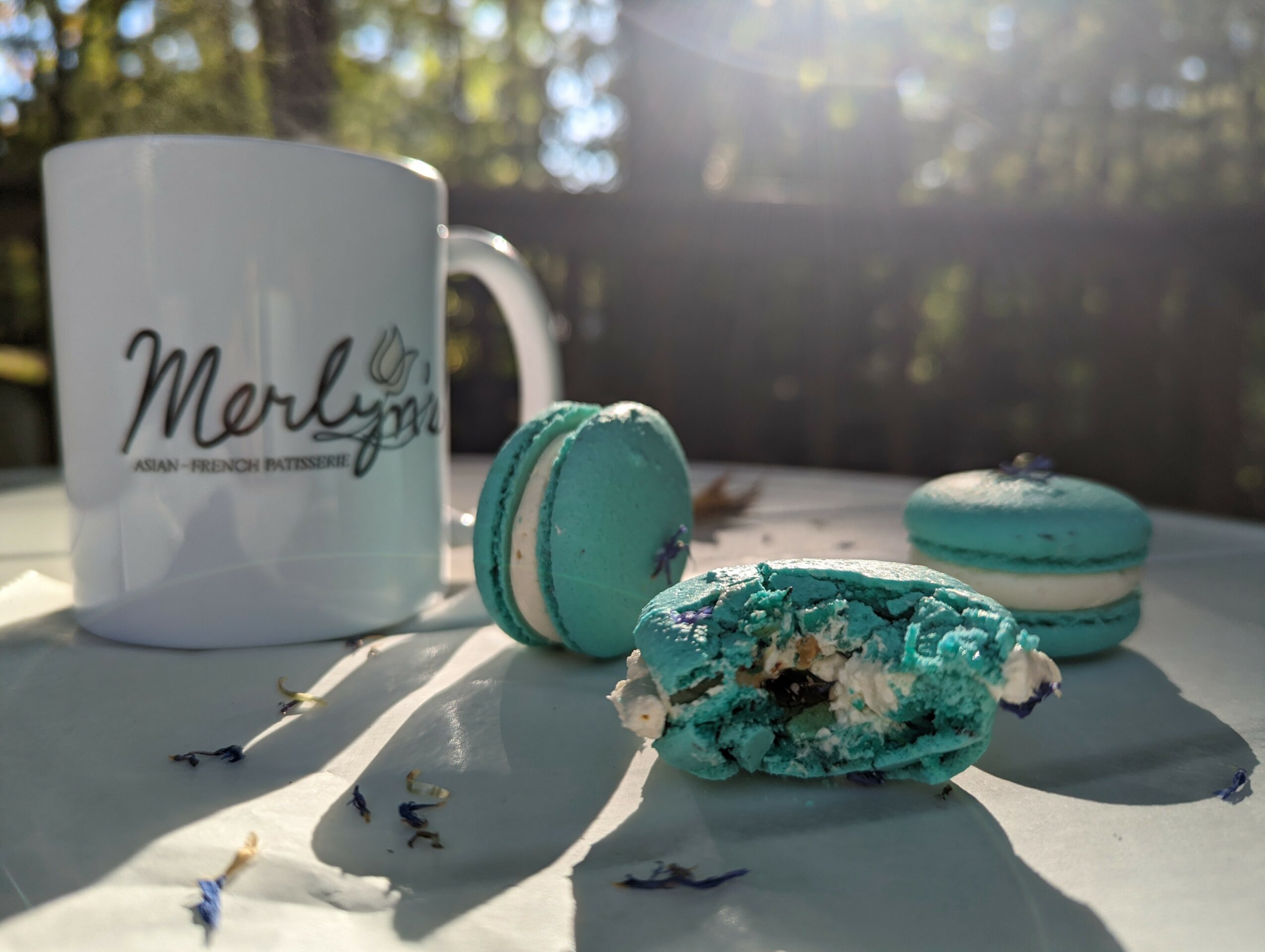 Three blue macarons and a mug with the word 'Merly' on a white surface outdoors, sunlight casting shadows.