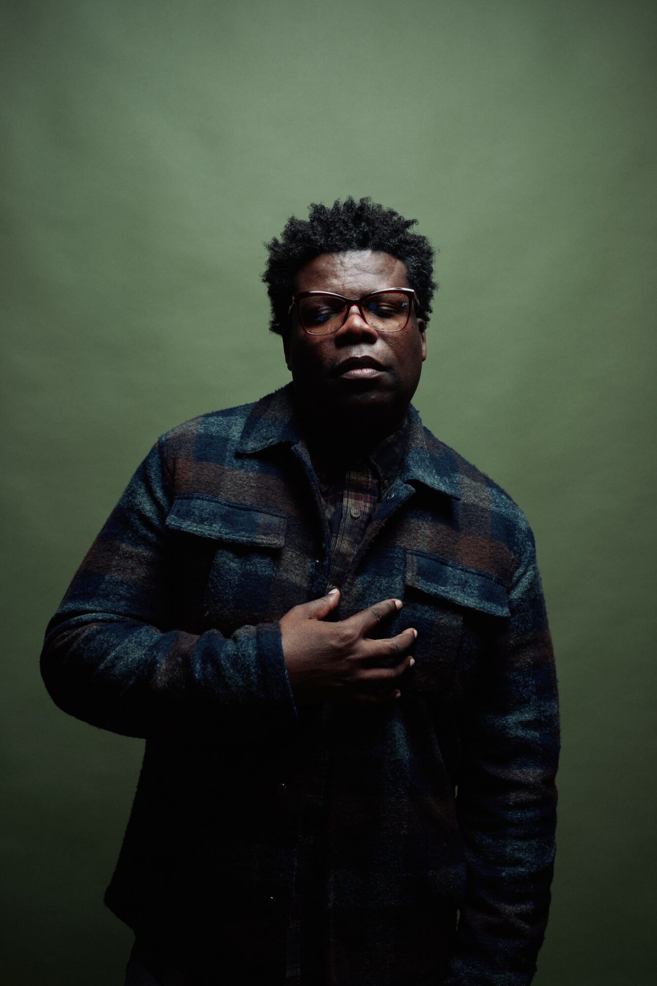 Person with glasses and curly hair wearing a plaid jacket, holding chest, against a green background.