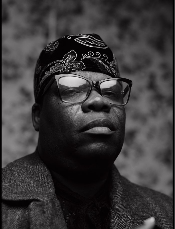 Portrait of a person wearing glasses, a bandana, and a coat, looking serious against a blurred background.