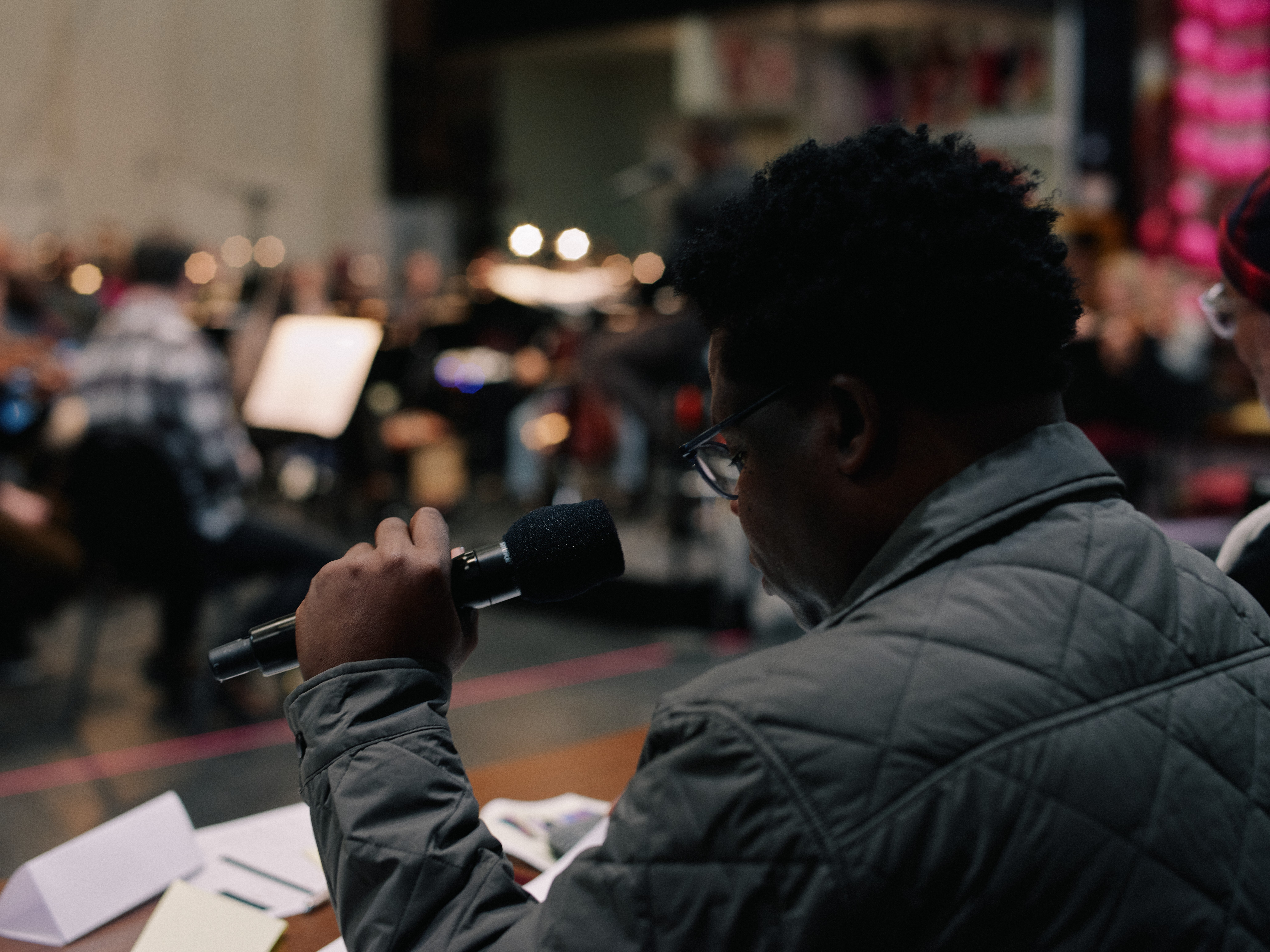 Person holding a microphone in a busy indoor setting with musicians in the background.
