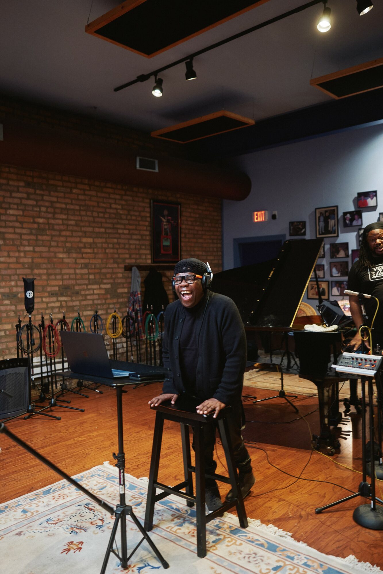 Person smiling, standing behind a music stand in a performance space with a brick wall and a piano in the background.