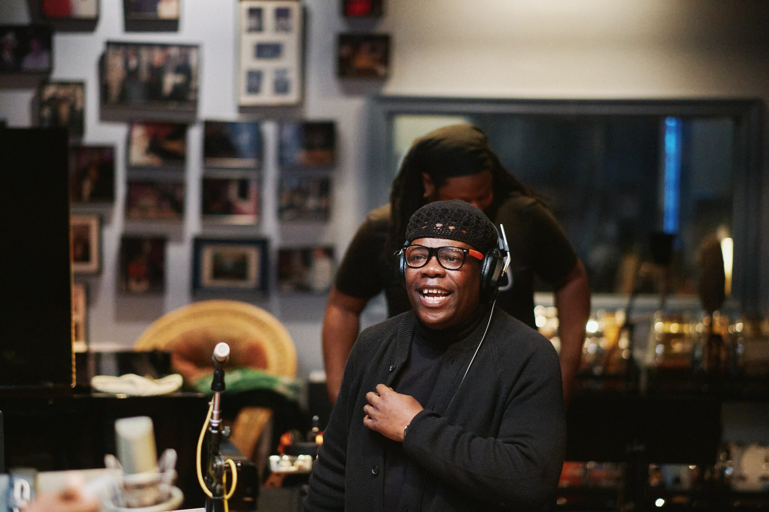 Person wearing headphones and glasses smiling, with a person behind in a room with photos and a window.