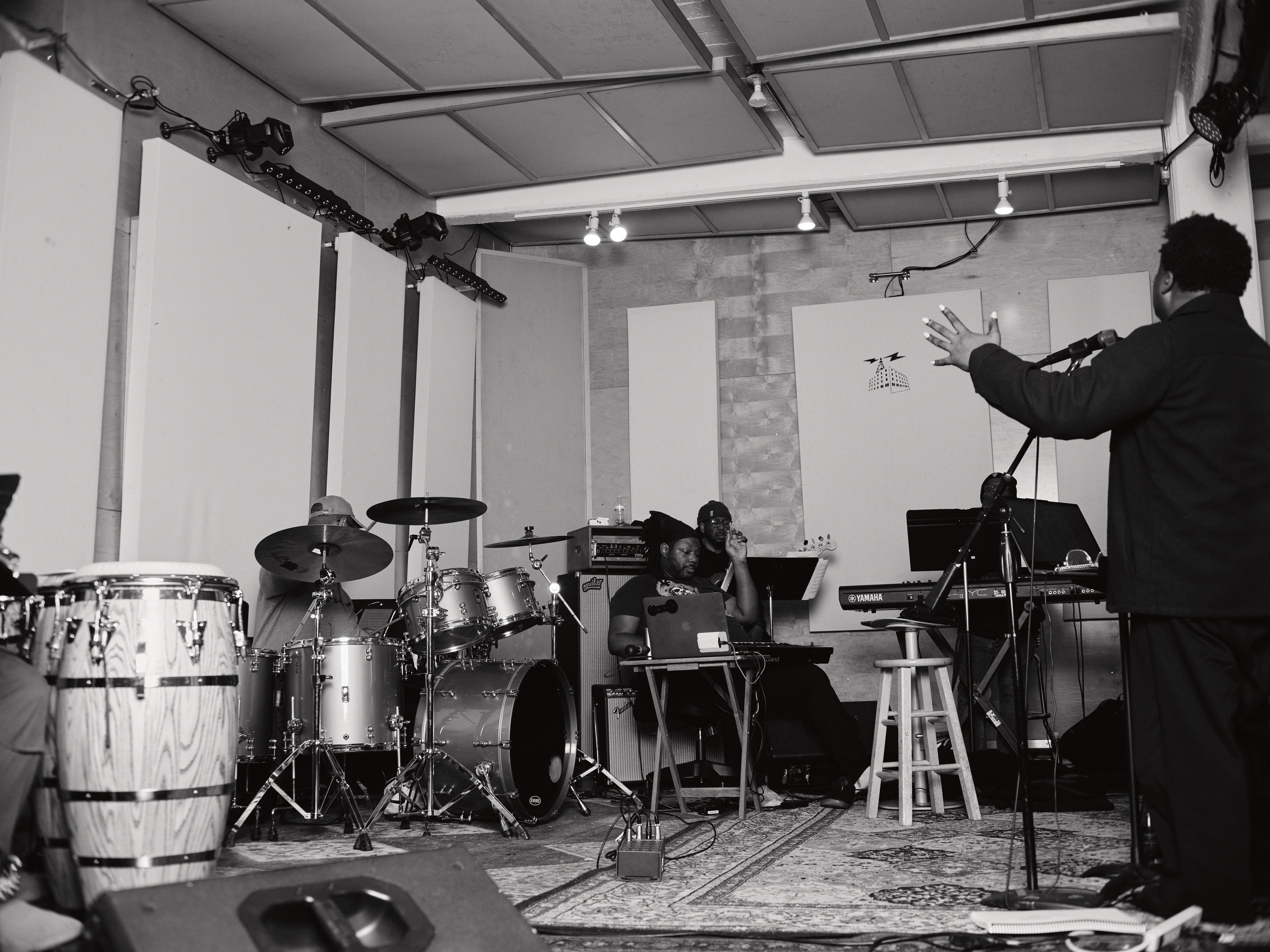Musicians perform on stage with drums, keyboard, and a person conducting or speaking, in a room with white walls and ceiling.