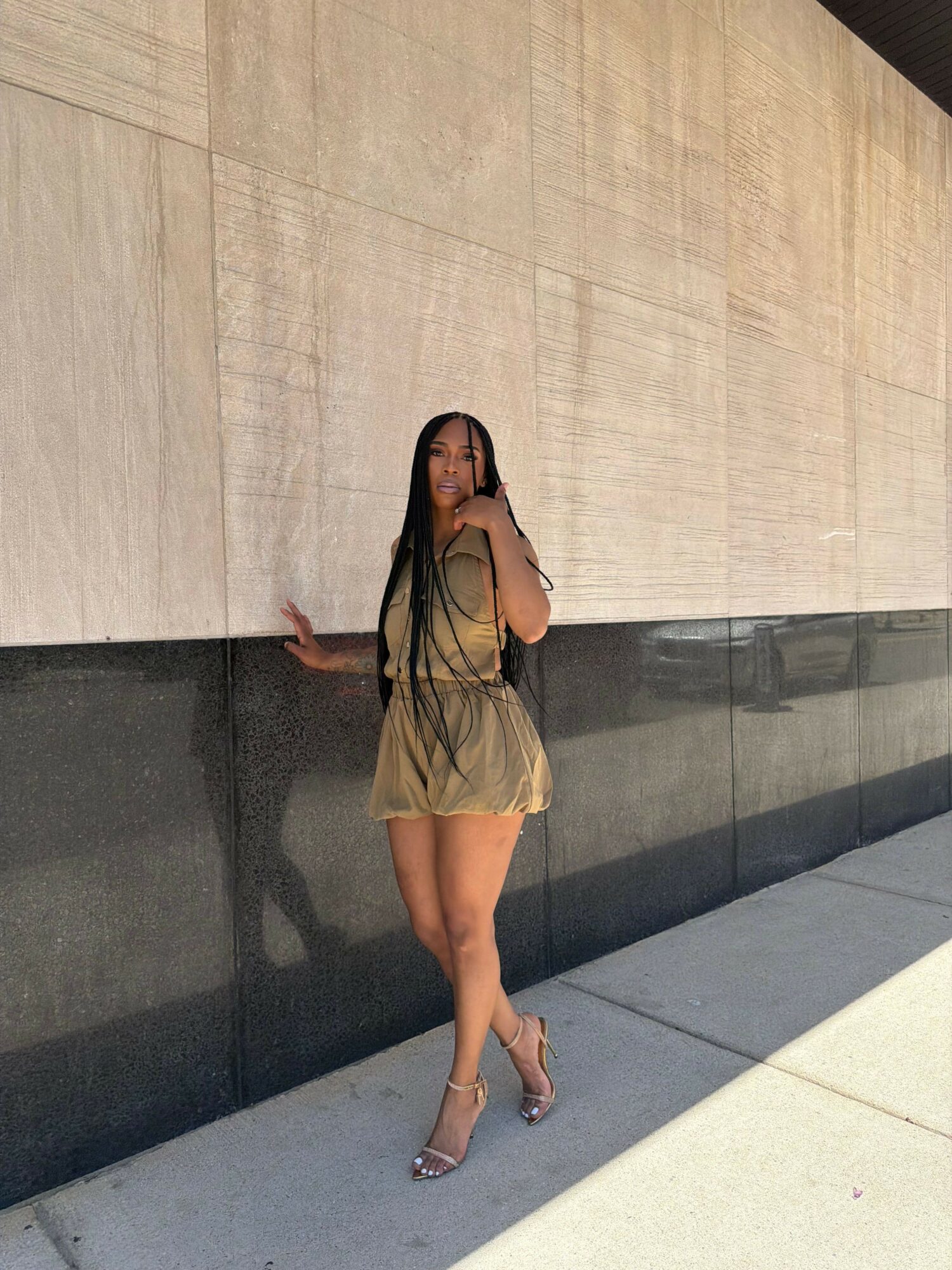 Woman in beige dress and heels leaning against a wall outdoors, with shadows cast on the ground.