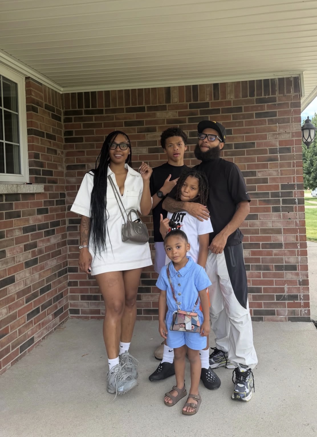 Four people stand outside a brick house, smiling, with a woman in white, three children, and a man in black.
