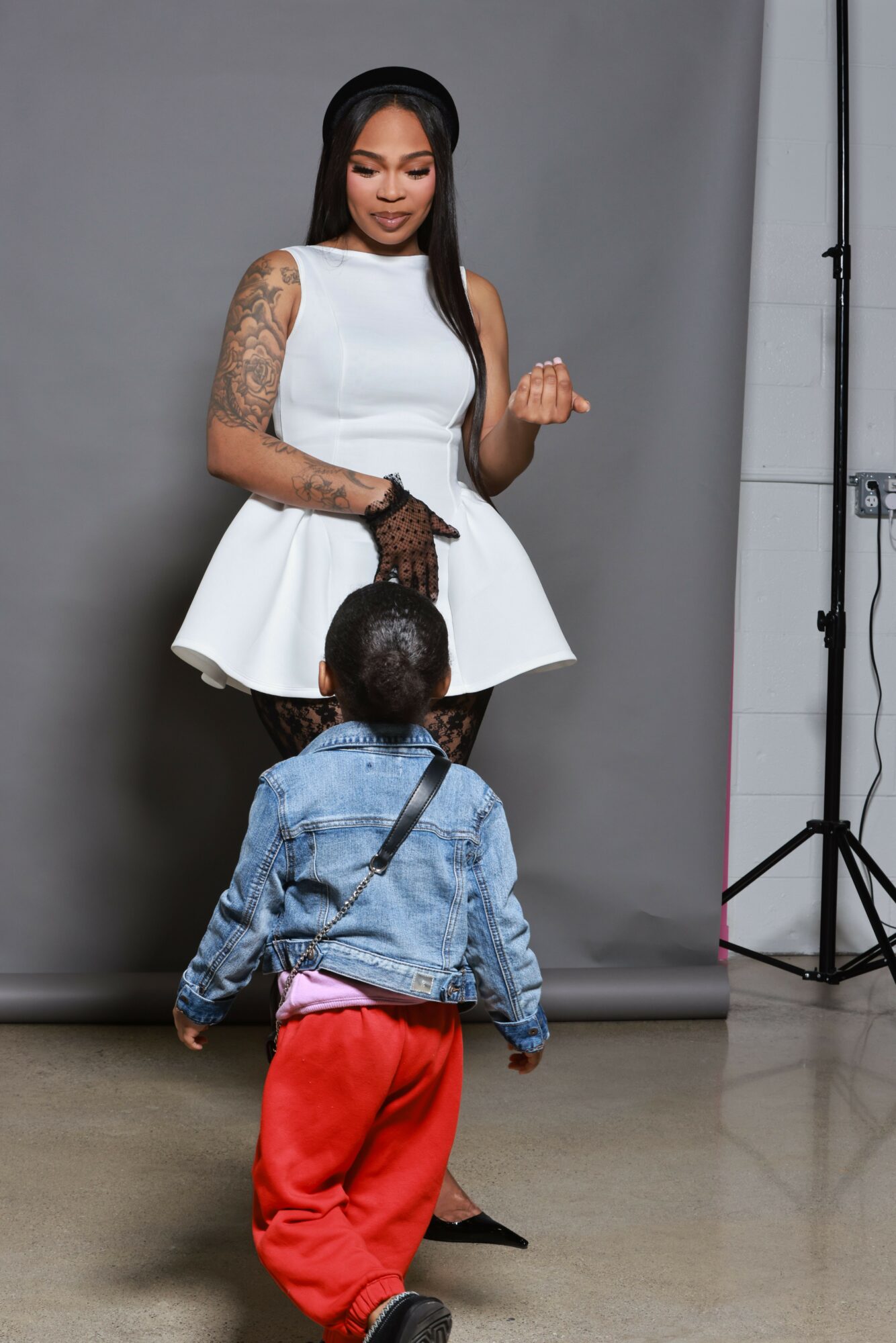 Woman with long black hair, white top, and tattoos, standing against a gray background, smiling and gesturing with her hand.