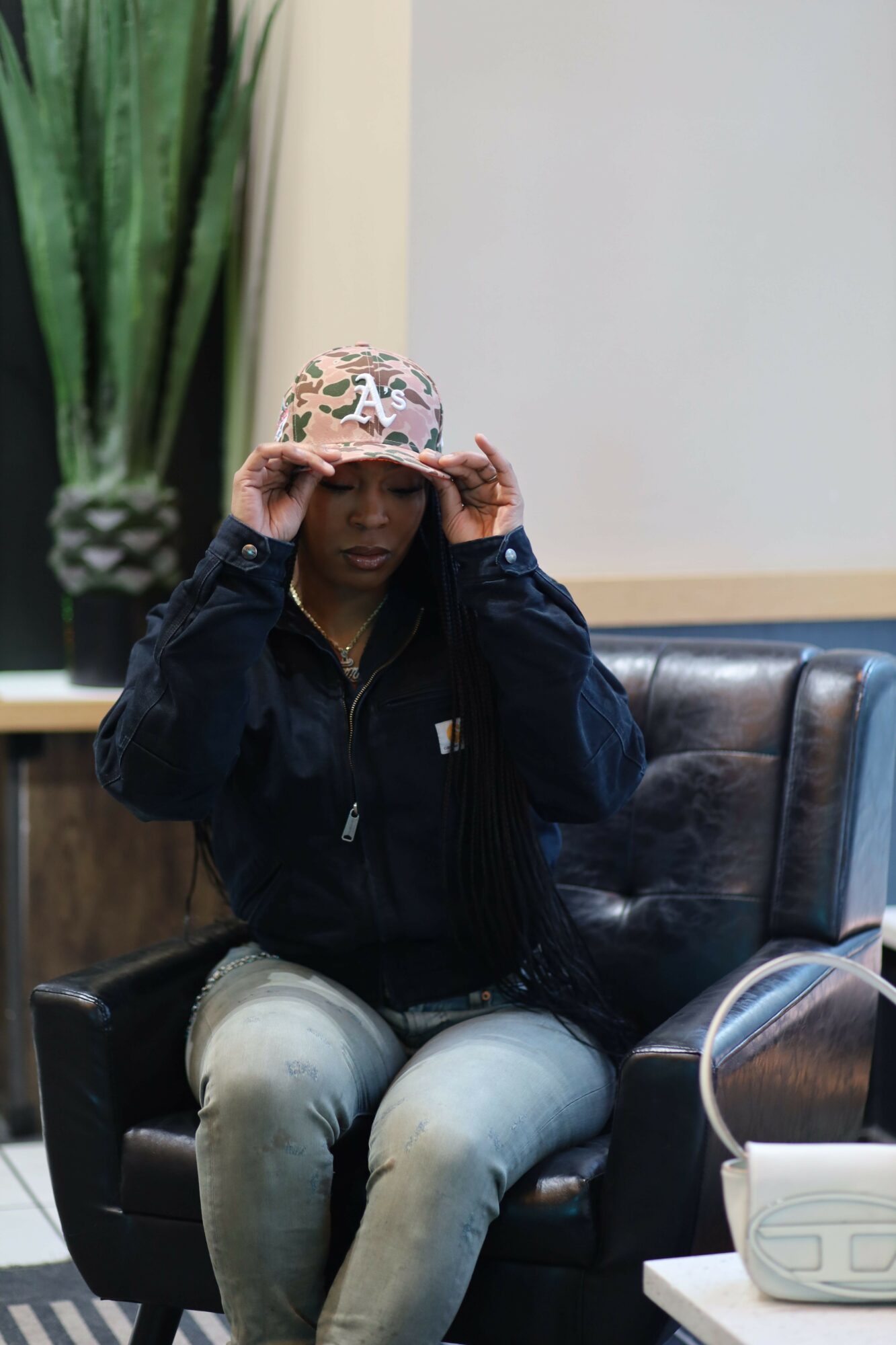 Person sitting on a black leather chair, wearing a patterned hat, dark jacket, and light pants, in a room with a white wall.