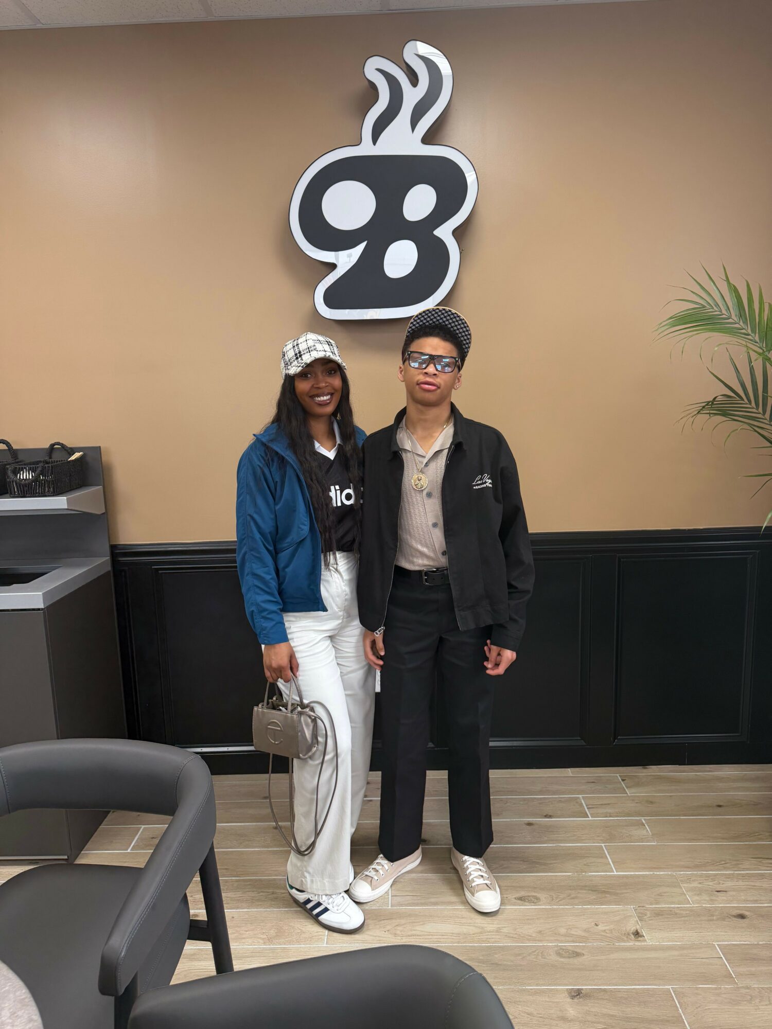 Two people standing indoors near a wall with a large decorative sign, one wearing glasses and a black jacket, the other in white pants and a blue jacket.