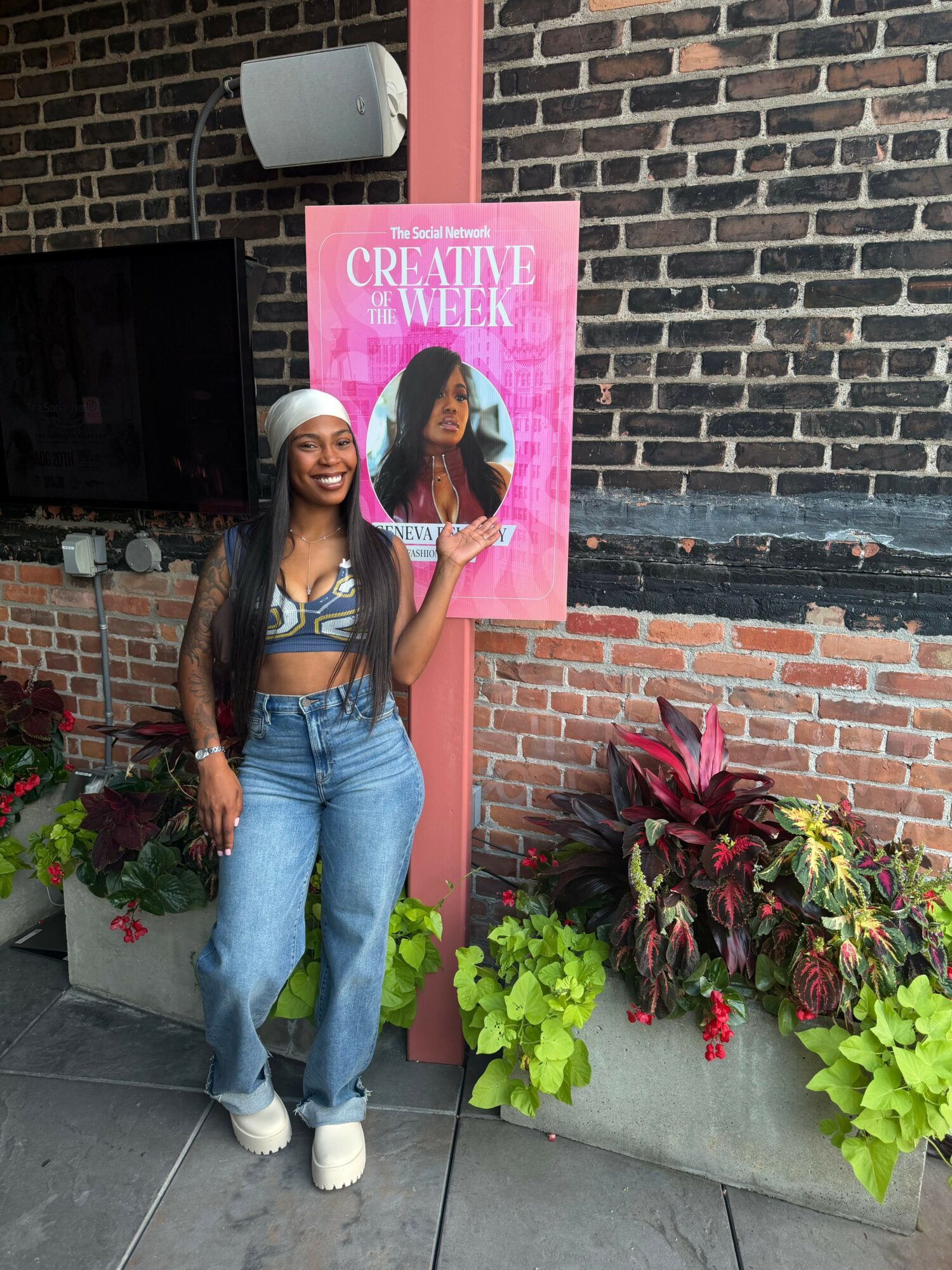 Woman standing next to a pink poster with a woman’s face, surrounded by potted plants and brick wall.