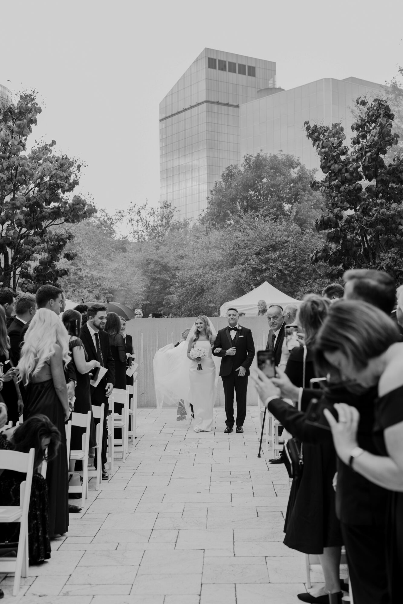 People gathered outdoors for a formal event, with a tall building and trees in the background.