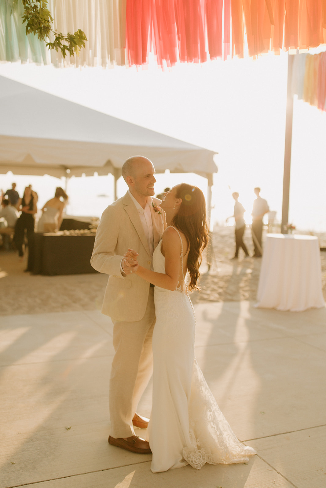 A couple dancing outdoors at a wedding reception, with a tent and guests in the background, under colorful hanging decorations.