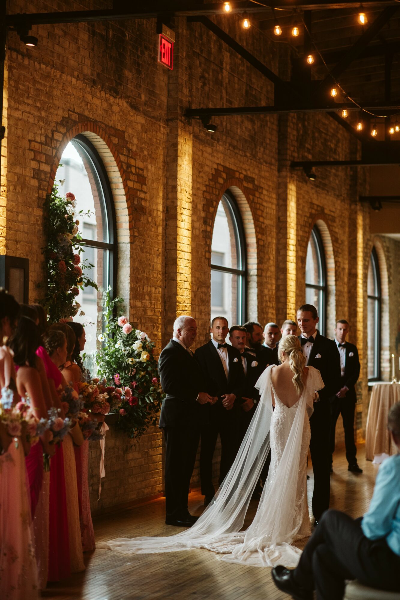 Wedding ceremony with bride and groom holding hands, guests, and brick walls with arched windows, warm lighting.