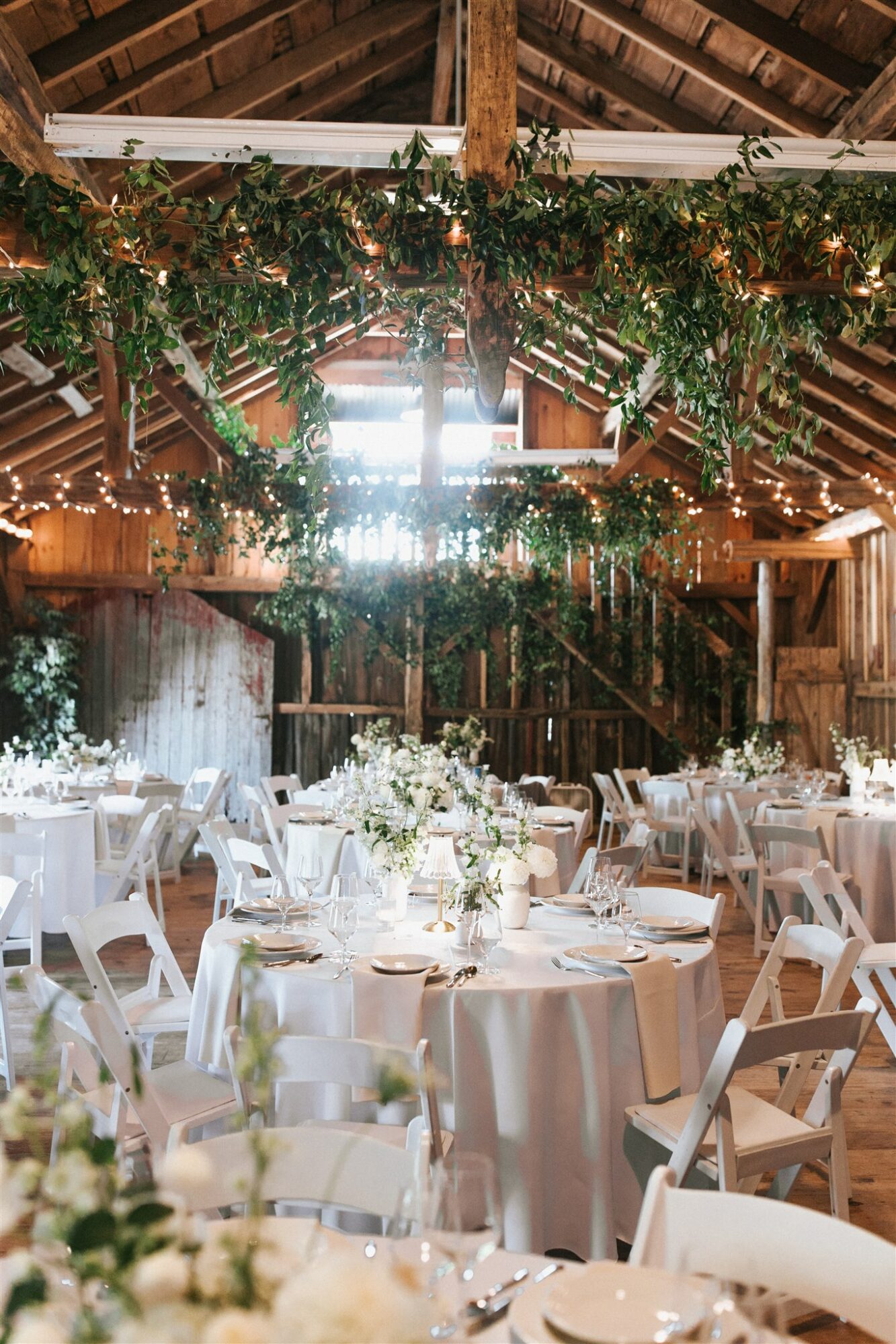 Wedding reception setup with round tables, white chairs, floral centerpieces, and hanging greenery inside a barn with wooden beams.