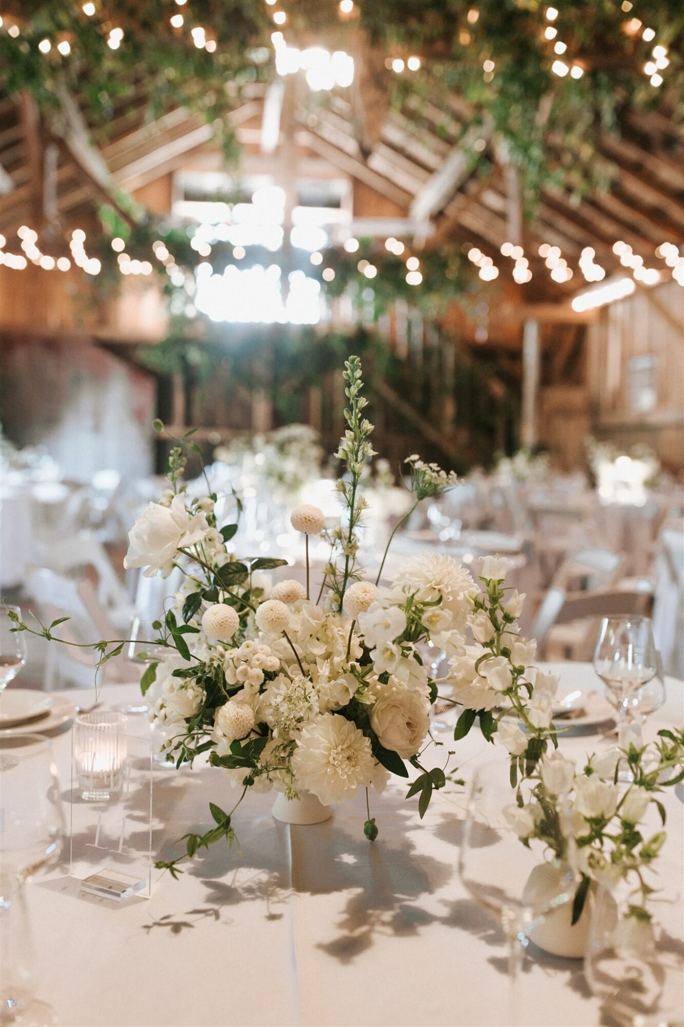 Elegant event table with a large floral centerpiece, surrounded by chairs and tableware, under string lights in a rustic venue.