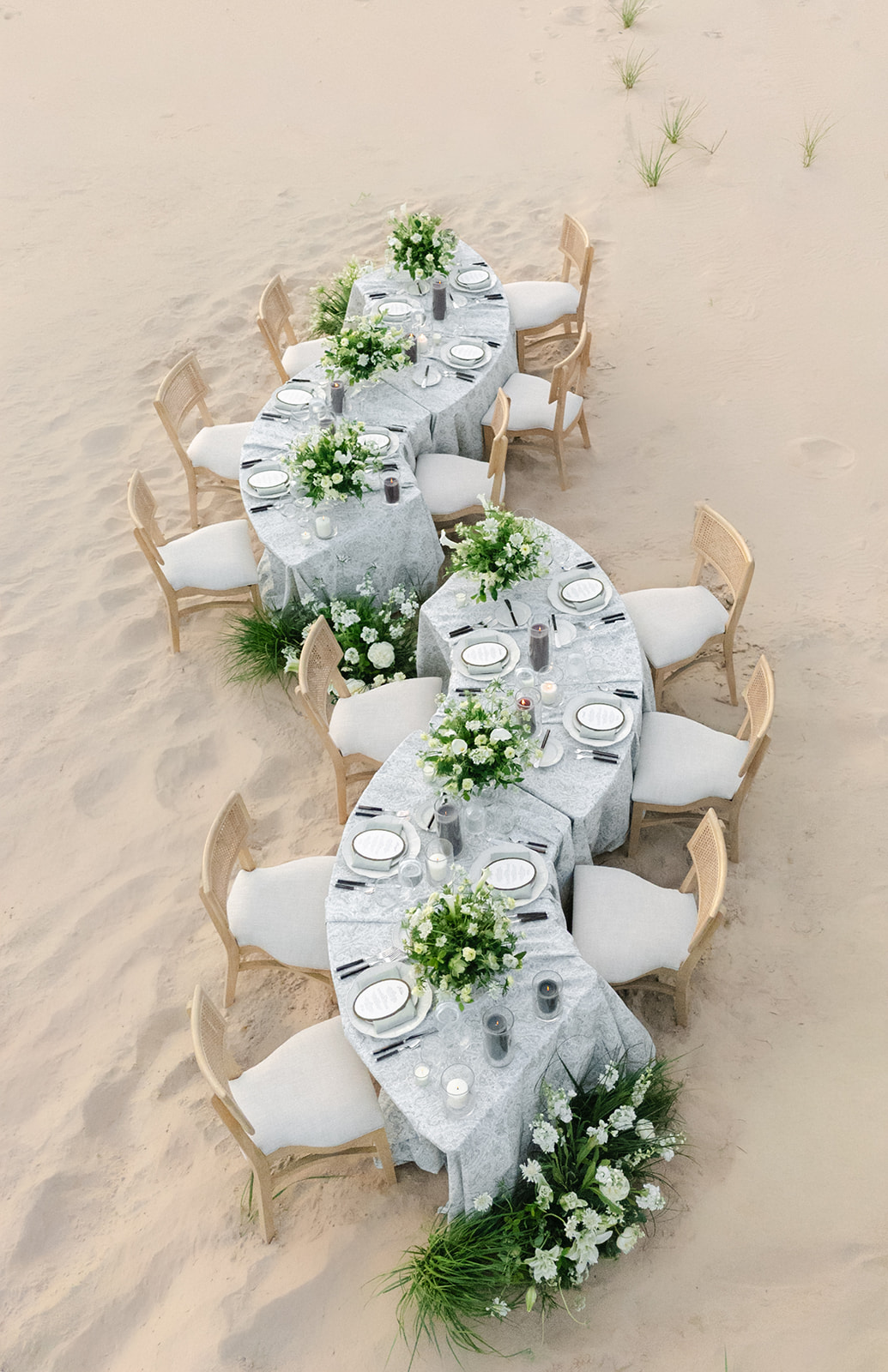 Long dining table set with white tablecloths, floral centerpieces, and place settings, on sandy ground with chairs around it.