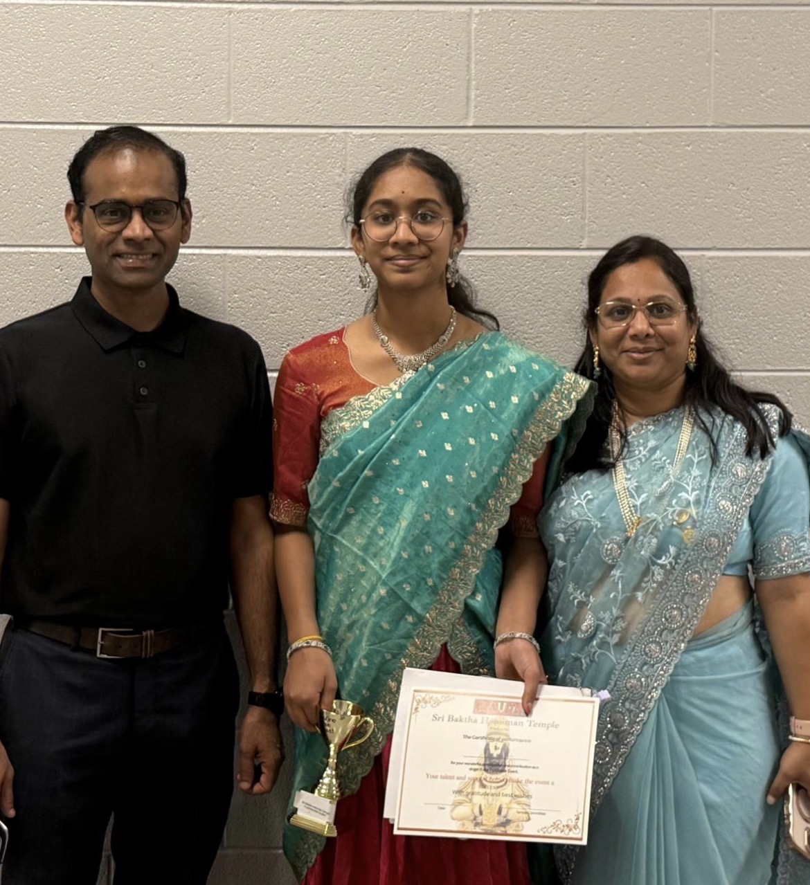 Three people standing against a plain wall, one holding a certificate and a trophy, all smiling.